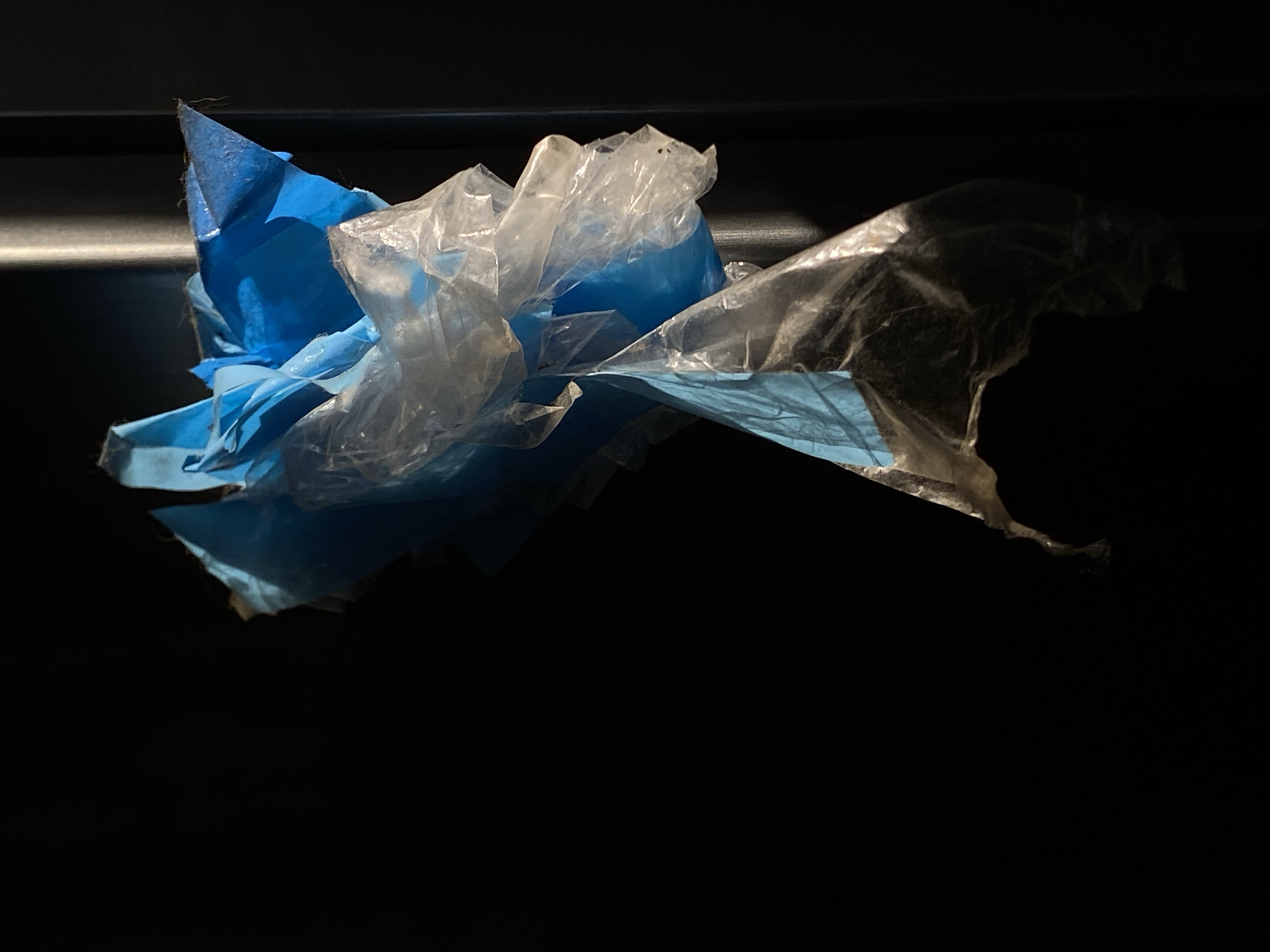In Belgrade, Serbia, a close-up shot focuses on a bundle of crumpled plastic bags, predominantly blue and translucent white, hanging against a dark, indistinct background. The lighting highlights the textures and translucent qualities of the plastic, creating dramatic shadows and highlights. The bags appear to be snagged or suspended, with some tearing and fraying visible along the edges. The scene is devoid of any human presence or activity, presenting a static still life of discarded materials.