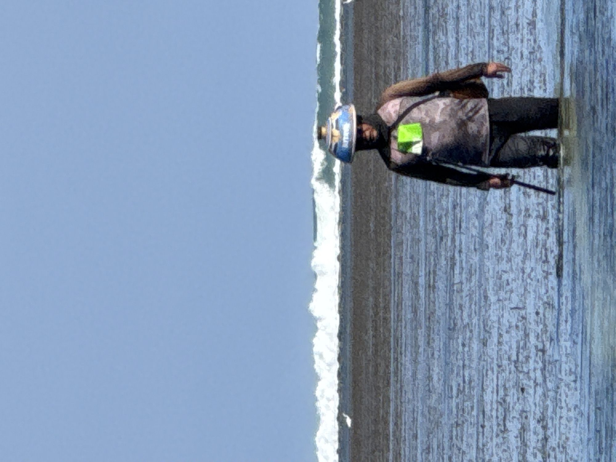 A person wearing a wide-brimmed hat stands in shallow water at the beach in Denpasar, Indonesia. The person is wearing a camouflage shirt with a green square design on the chest and dark pants. They are holding a fishing rod in their right hand and have a bag slung over their left shoulder. The water is calm, and a wave is breaking in the distance. The sky is clear and blue.