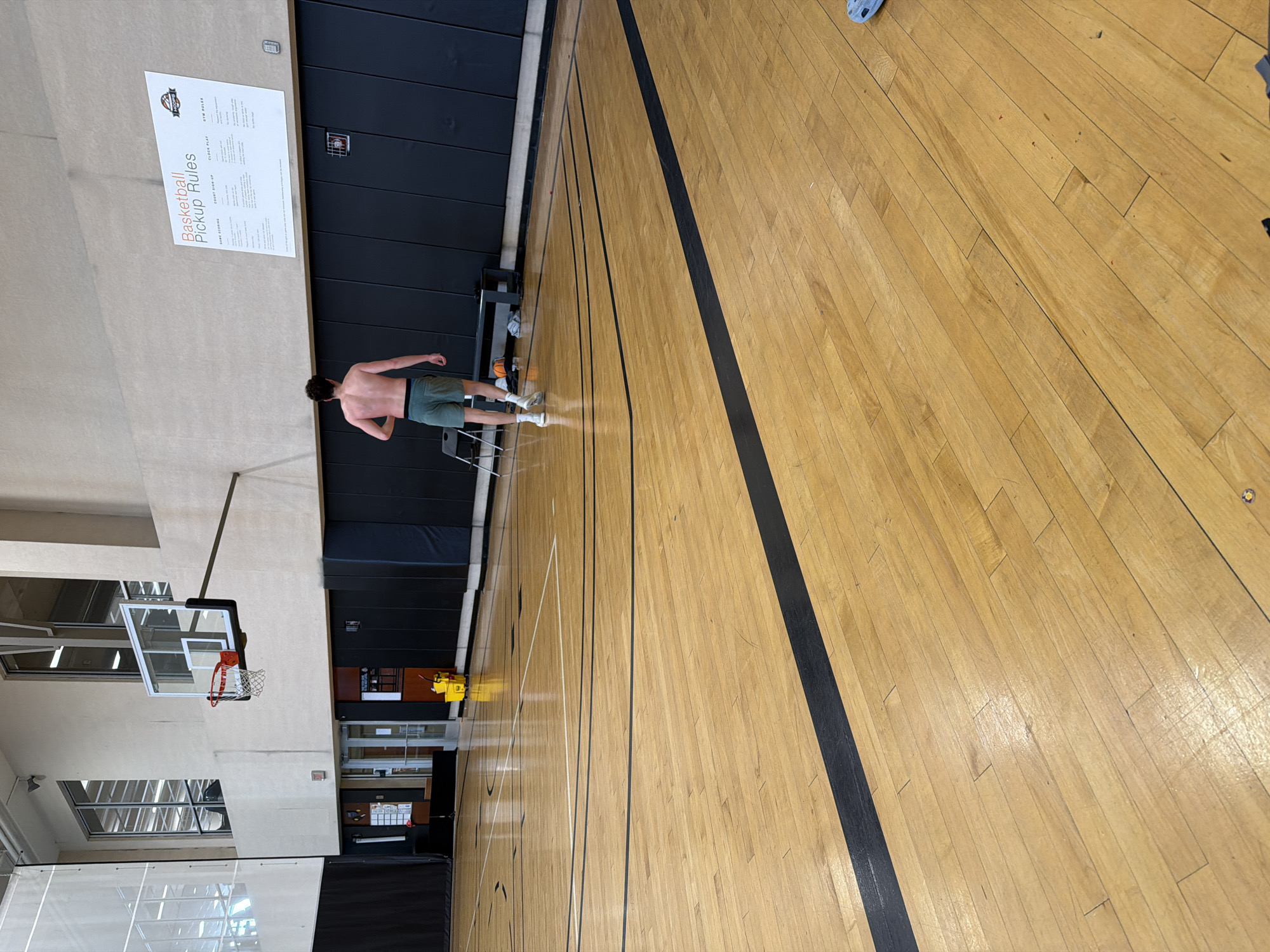 An indoor basketball court in Beachwood, United States of America, is depicted from a slightly elevated, rear-facing perspective. The foreground features a light-toned wooden court floor marked with black and white lines. The floor shows a reflective finish and minor scuffing.
The primary subject is a male individual, seen from behind, walking away from the camera. He is shirtless, has dark, curly hair, and wears dark green athletic shorts with a black waistband, white socks, and white sneakers. He is moving towards the far end of the court, slightly to the right of the central axis.
On the left side of the image, a basketball hoop with a clear backboard, white frame, red rim, and white net is affixed to a light beige upper wall. Below the hoop, the lower wall is covered in dark gray padded panels. Above the padded wall, two large windows reveal an interior area of the building. To the left of the hoop and padded wall, a set of double glass doors is visible, with a yellow mop bucket and red mop handle positioned on the court floor nearby.
On the right side of the image, a large white sign titled "Basketball Pickup Rules" is mounted on the light beige upper wall. The sign is divided into sections including "GAME SCORING," "COURT SIGN-UP," "CLOCK PLAY," and "GYM RULES," and features a "MOORE" logo. Below the sign, the lower wall section is also covered in dark gray padded panels. A dark, low bench or table is placed against this padded wall, with a single orange basketball resting on the floor in front of it. A dark chair is also present to the left of the walking individual. The gymnasium is illuminated by overhead artificial lighting.