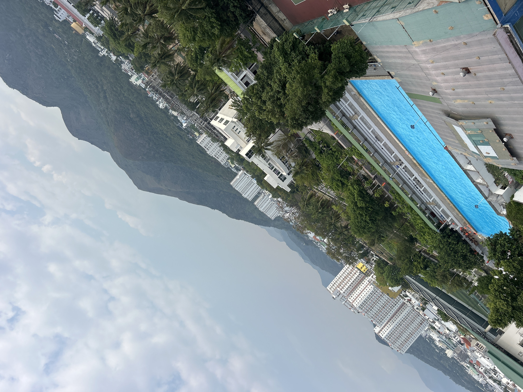 The image depicts an aerial view of a cityscape in Bac Nha Trang Ward, Vietnam, adjacent to a mountain range. The foreground features a rectangular, blue swimming pool alongside a building complex with a light gray roof. Dense green trees surround the pool and buildings. Further in the distance, white high-rise buildings form a distinct urban landscape along the coastal stretch. The sky is overcast with visible cloud formations. The mountain range is characterized by its green slopes and ridgelines.