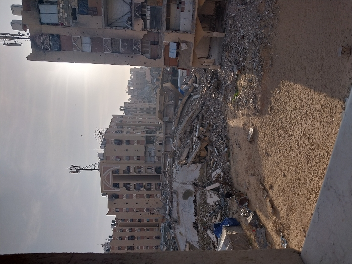 This image captures a wide-angle view of extensive urban devastation in Khan Yunis, Palestinian Territory, likely observed from an elevated position within a structure, evidenced by a concrete ledge in the lower foreground. The scene is dominated by a large expanse of sandy ground and massive piles of building rubble in the midground, consisting of broken concrete slabs, twisted rebar, and assorted debris, suggesting complete structural collapse of multiple buildings. In the background, several multi-story residential buildings, primarily light brown or tan, exhibit significant damage: facades are scarred with numerous impact holes, windows are blown out or covered, and sections appear charred or partially collapsed. Communications towers with antennae are visible on the rooftops of some of these damaged structures. A particularly heavily damaged building stands prominently on the right, its entire facade pockmarked with numerous small holes, with exposed interiors and remnants of window frames. The sun, low in the sky, is visible through a hazy atmosphere, casting a strong glare from behind the damaged buildings on the right. No people or discernible actions are visible in the scene.