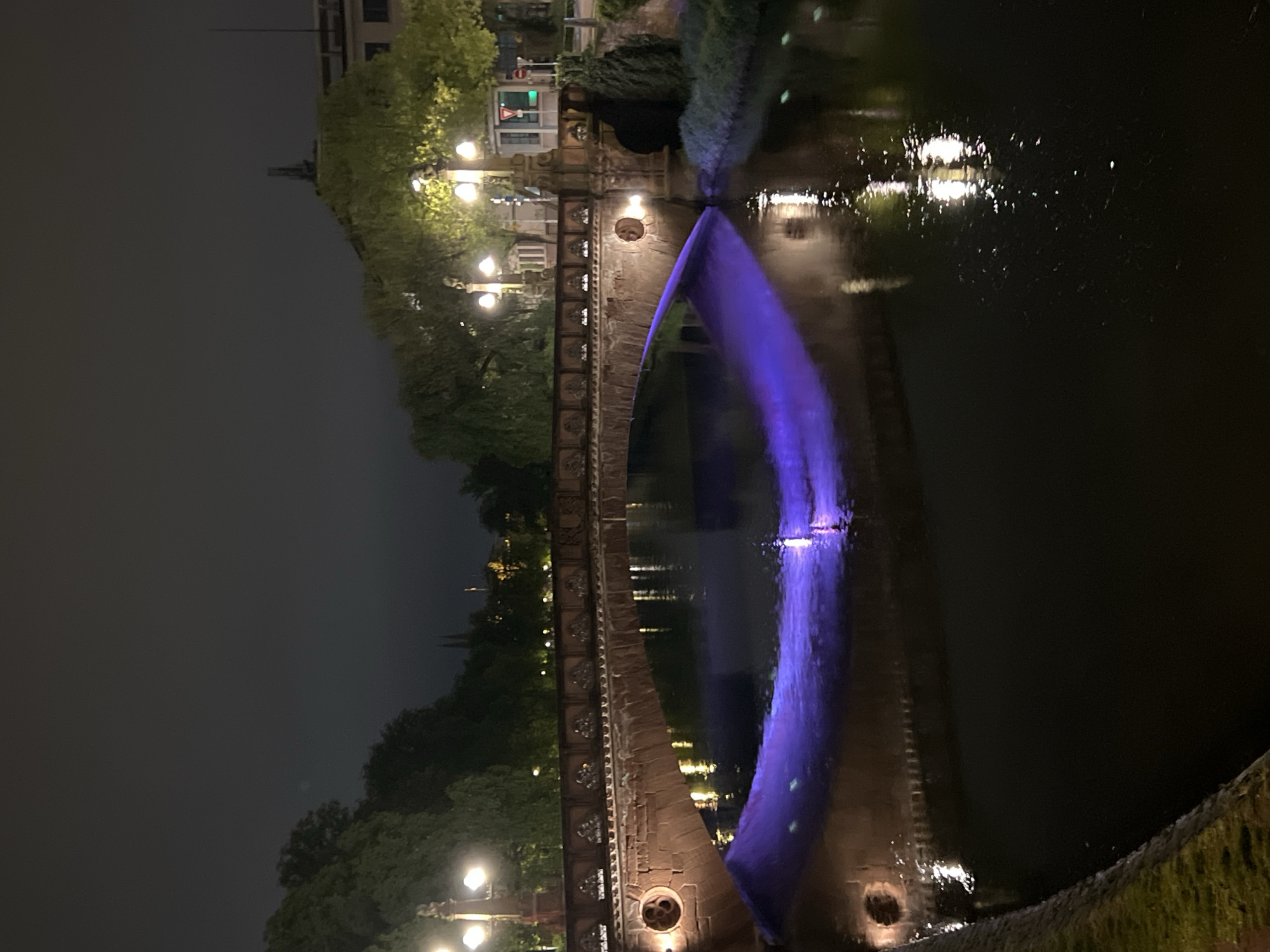 The image shows the Ponts Couverts in Strasbourg, France at night. The bridge is made of stone and has several arches. A blue light illuminates the water flowing through one of the arches. Trees line the canal and lights illuminate the area. The sky is dark, suggesting it is nighttime. The canal water is dark and reflective.