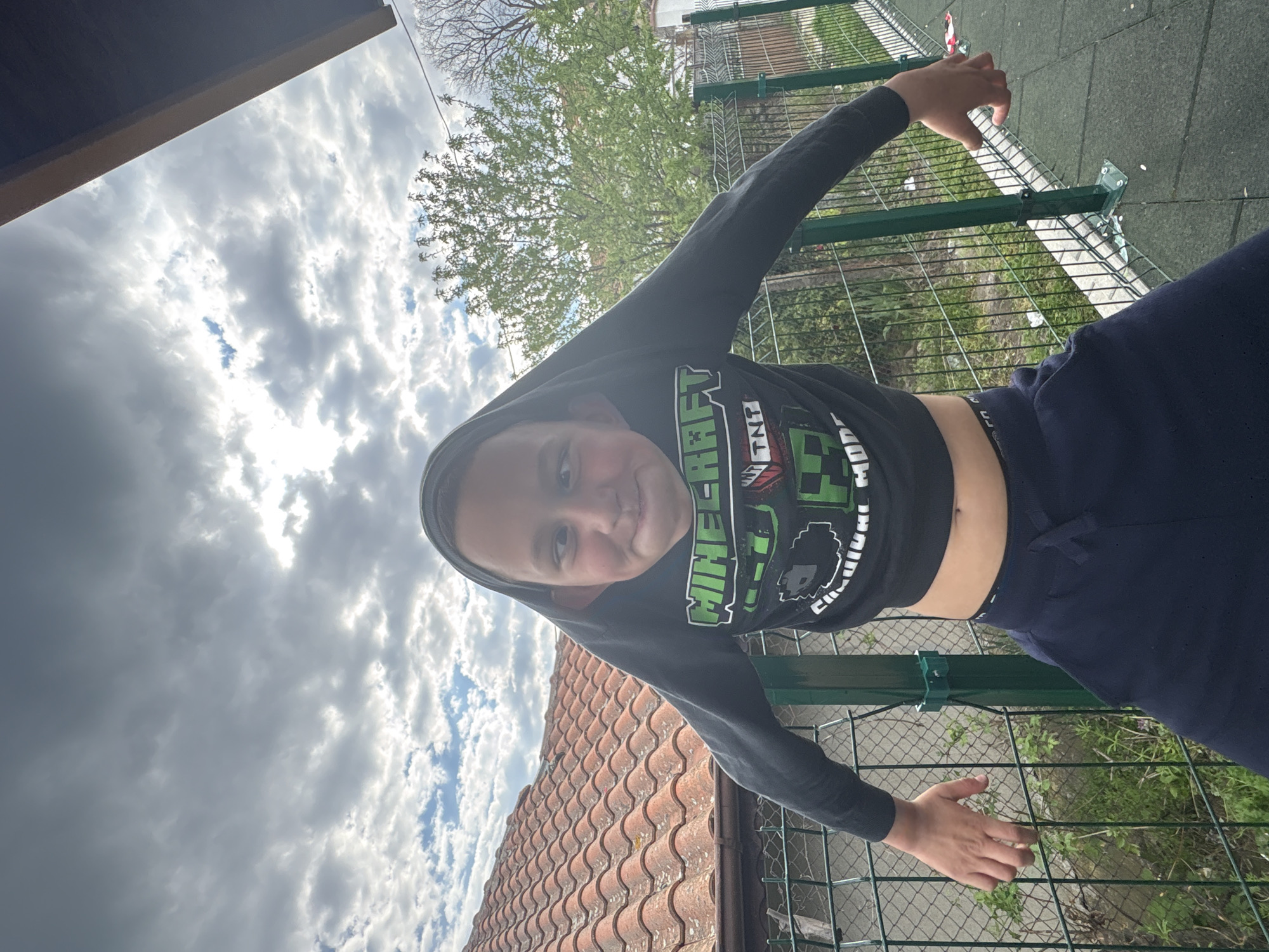A young person wearing a Minecraft hooded shirt and navy sweatpants is positioned between a building with a red-tiled roof and a green wire fence. The background features a sky with cloudy weather. The location is in the city of Unknown, Unknown.
