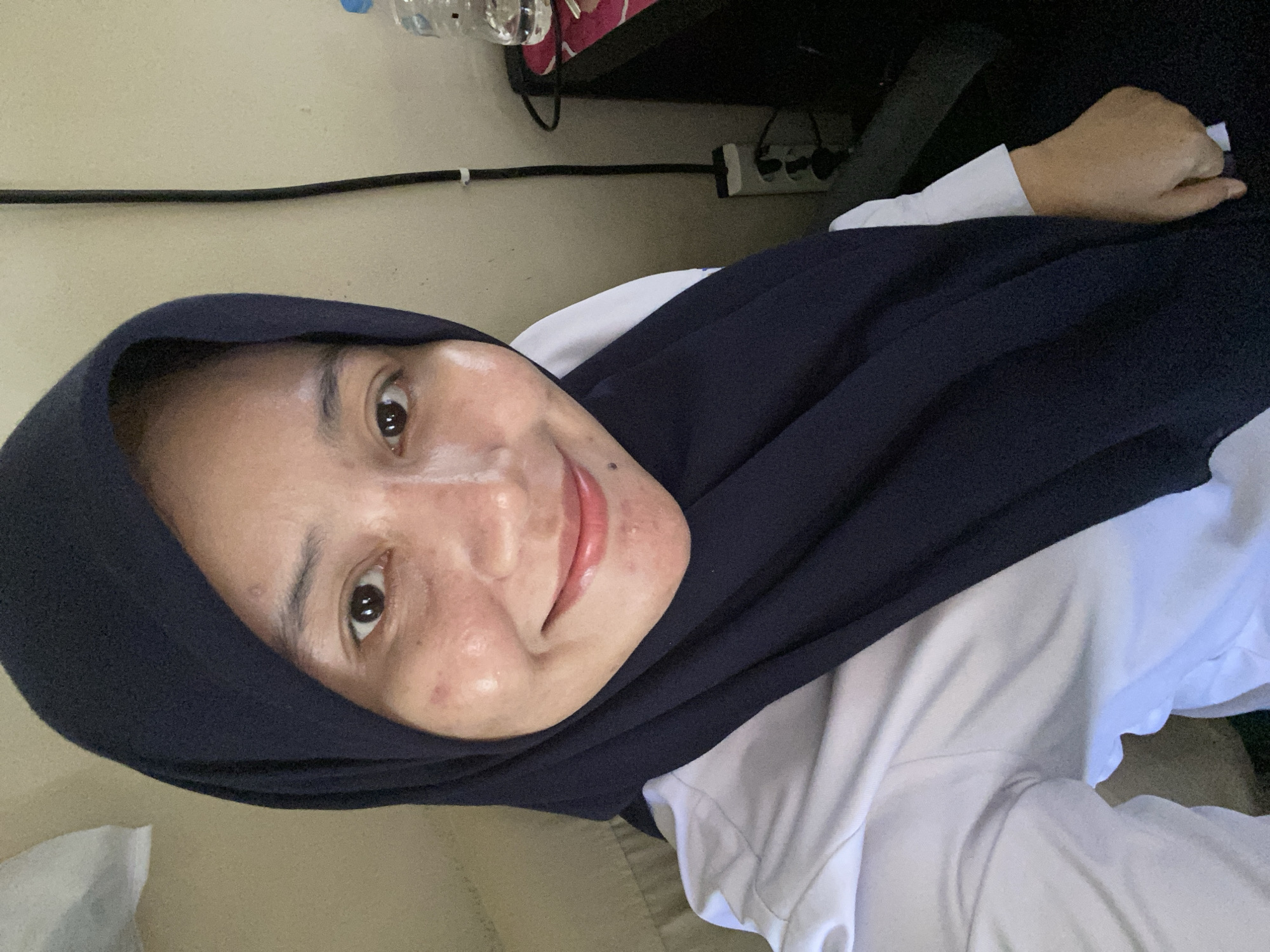 A medium shot selfie of a woman wearing a dark blue hijab and a white collared shirt is captured in a seated position. The woman has a fair complexion with visible acne scars and active blemishes on her cheeks and forehead. Her eyes are dark brown and she is smiling faintly, with a subtle glint of moisture on her lips. A black cable hangs vertically on the beige wall behind her, connecting to a power strip with multiple outlets. To the right of the woman, a clear plastic water bottle sits on a dark surface. The background suggests an indoor setting, possibly an office or a room in Banjarbaru, Indonesia, indicated by the presence of office-like furniture and electrical outlets.