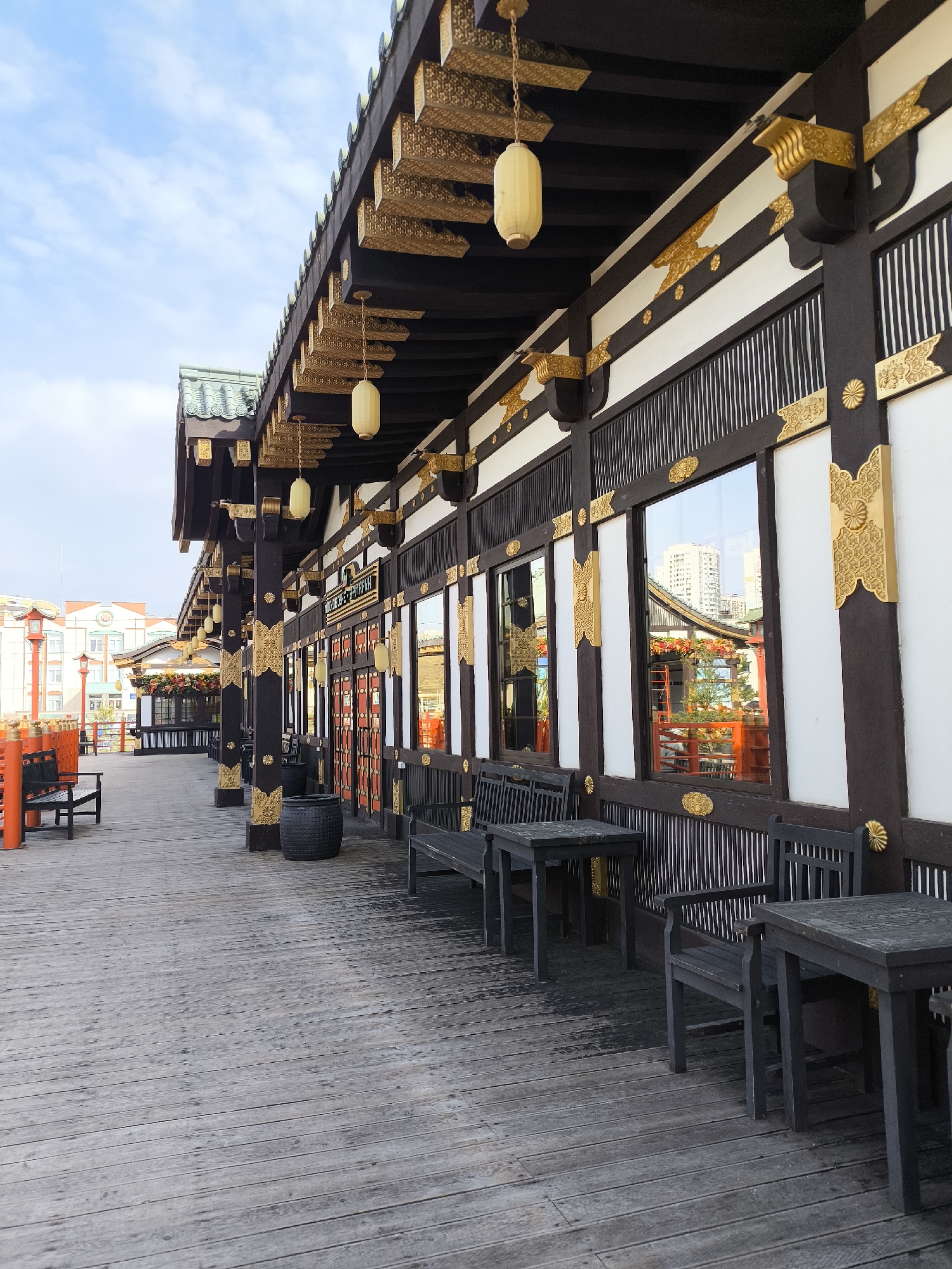 The image depicts a covered wooden walkway alongside a building with East Asian architectural details, likely in a public space in Moscow, Russia. The building has a dark-colored wooden frame with white panels, adorned with gold ornamentation. Large windows reflect nearby buildings. Hanging lanterns are suspended from the eaves. Dark wooden benches and tables are situated along the walkway. The walkway itself is constructed of weathered wooden planks. The sky is overcast with some patches of blue. In the background, a red building can be seen, along with glimpses of other city buildings. There are no people immediately visible in the foreground.