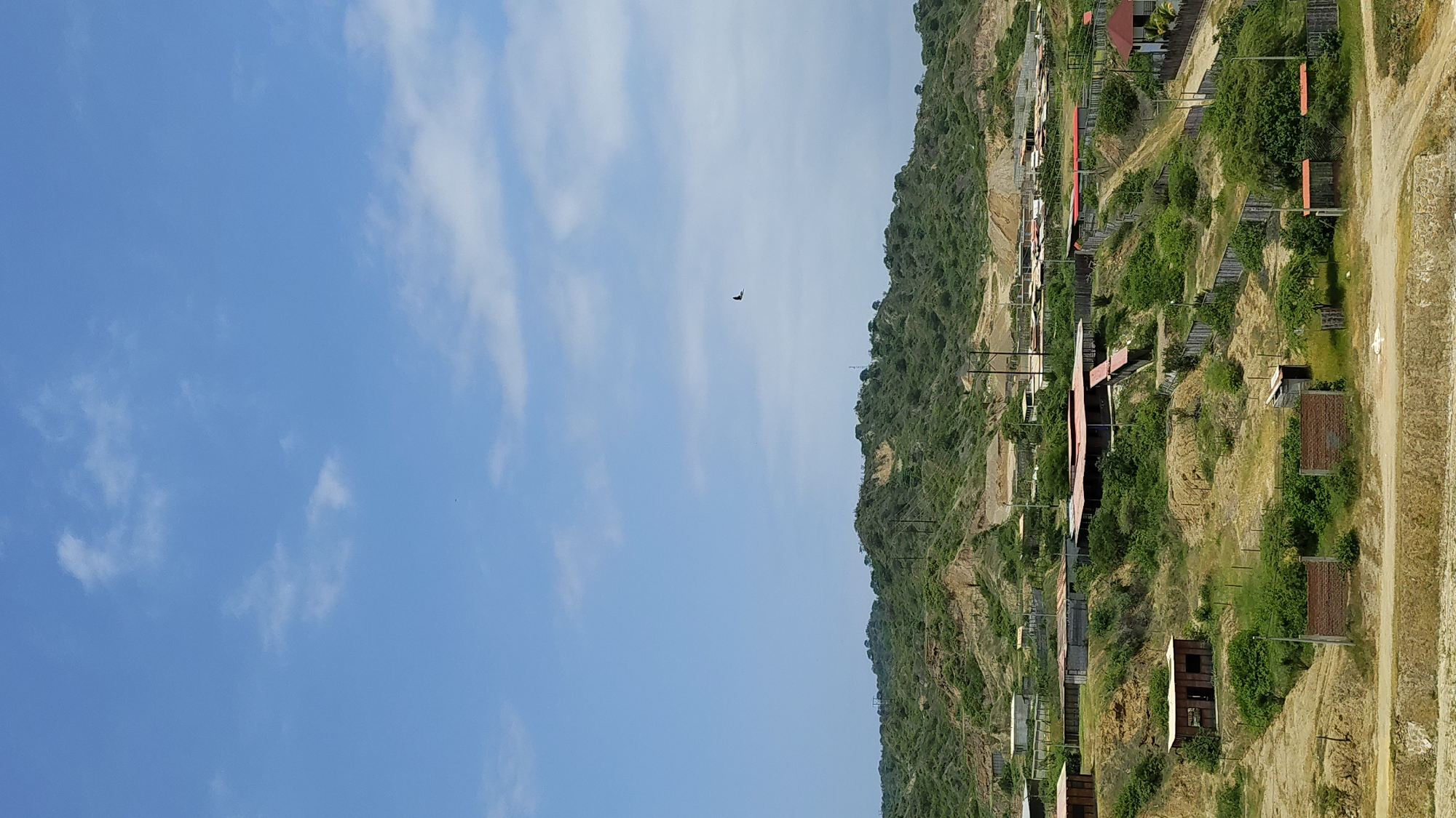The image depicts a daytime scene in Bocapán, Peru. The right side of the frame shows a hillside covered in green vegetation and a series of small structures, some with red roofs. A dirt road runs along the base of the hill. The left side of the frame is dominated by a bright blue sky with scattered white clouds. A single bird is visible in flight against the sky. The image is rotated counter-clockwise by approximately 90 degrees.