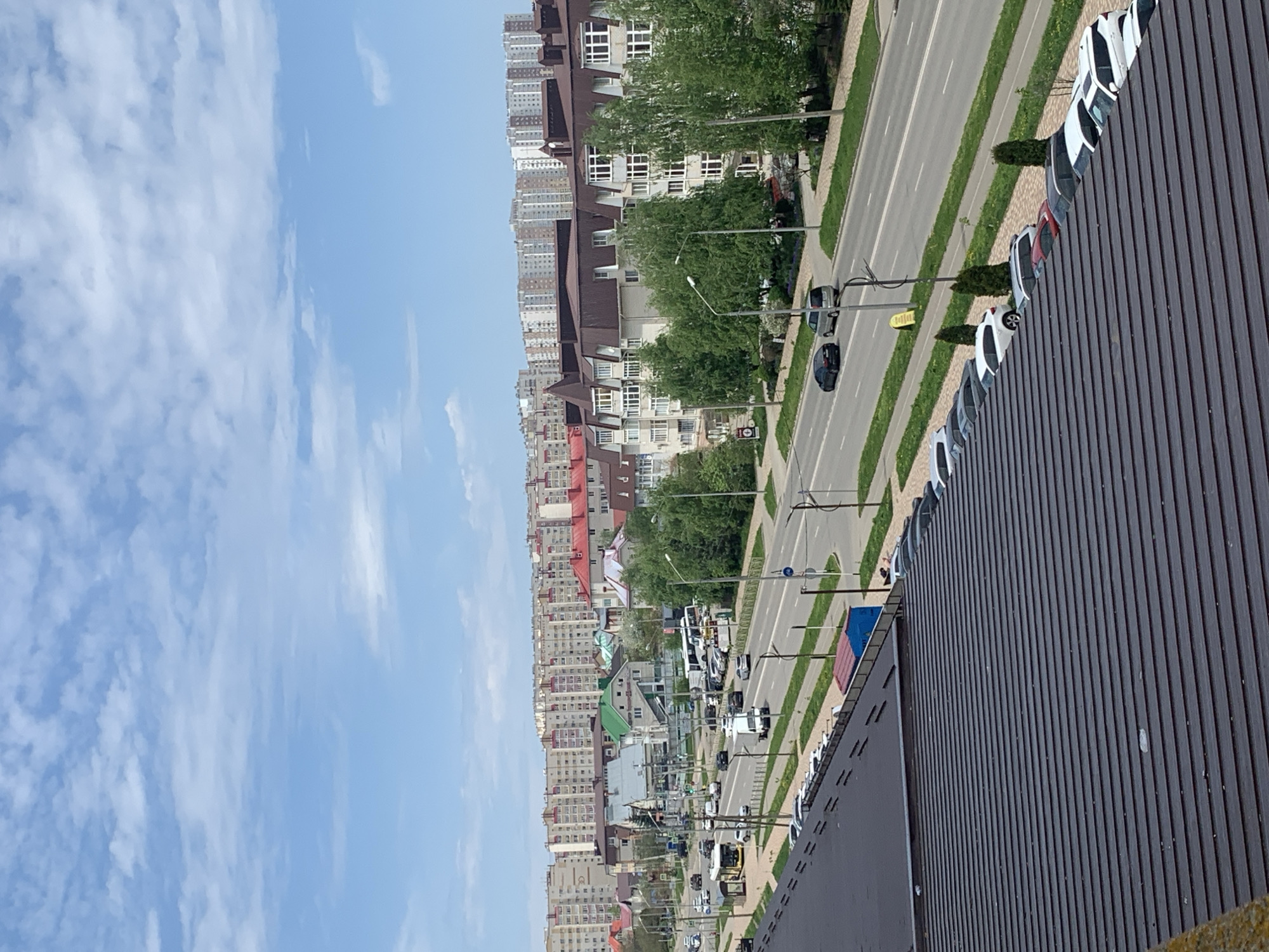Here is a description of the image:
The image depicts an aerial view of a city street. On the left side of the frame is a clear blue sky with scattered white clouds. In the middle of the image, along the horizon line, are several buildings of varying heights and architectural styles. The buildings are mostly multi-story residential structures with a mix of colors and materials.
A multi-lane road runs alongside the buildings, with a median strip separating the lanes. Cars and trucks are visible on the road, moving in both directions. Streetlights and trees line the sidewalks on either side of the road.
In the foreground on the right, the image shows a dark, corrugated metal surface, suggesting the view is from a rooftop or high vantage point. A row of parked cars is also visible along the edge of this surface.
The city is in Unknown, Unknown. The overall scene appears to be a normal day in a bustling urban environment.