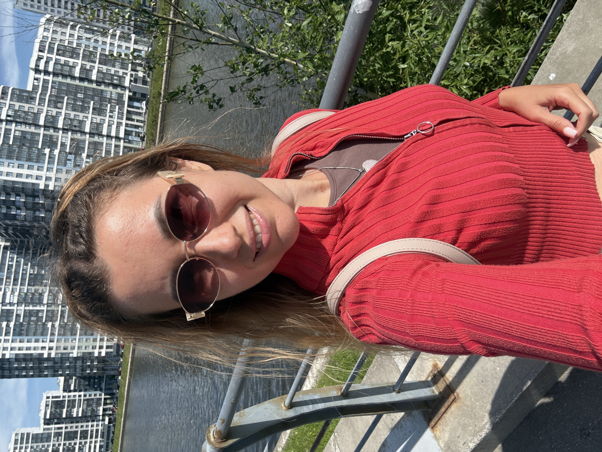 A selfie is taken by a Caucasian woman with blonde hair pulled back, wearing sunglasses, a red zip-up sweater, and a grey shirt underneath. She is smiling and looking at the camera. In the background, there is a body of water, a metal railing, and several modern high-rise apartment buildings. Green trees and foliage are also visible to the right of the woman. The image appears to be taken on a sunny day.