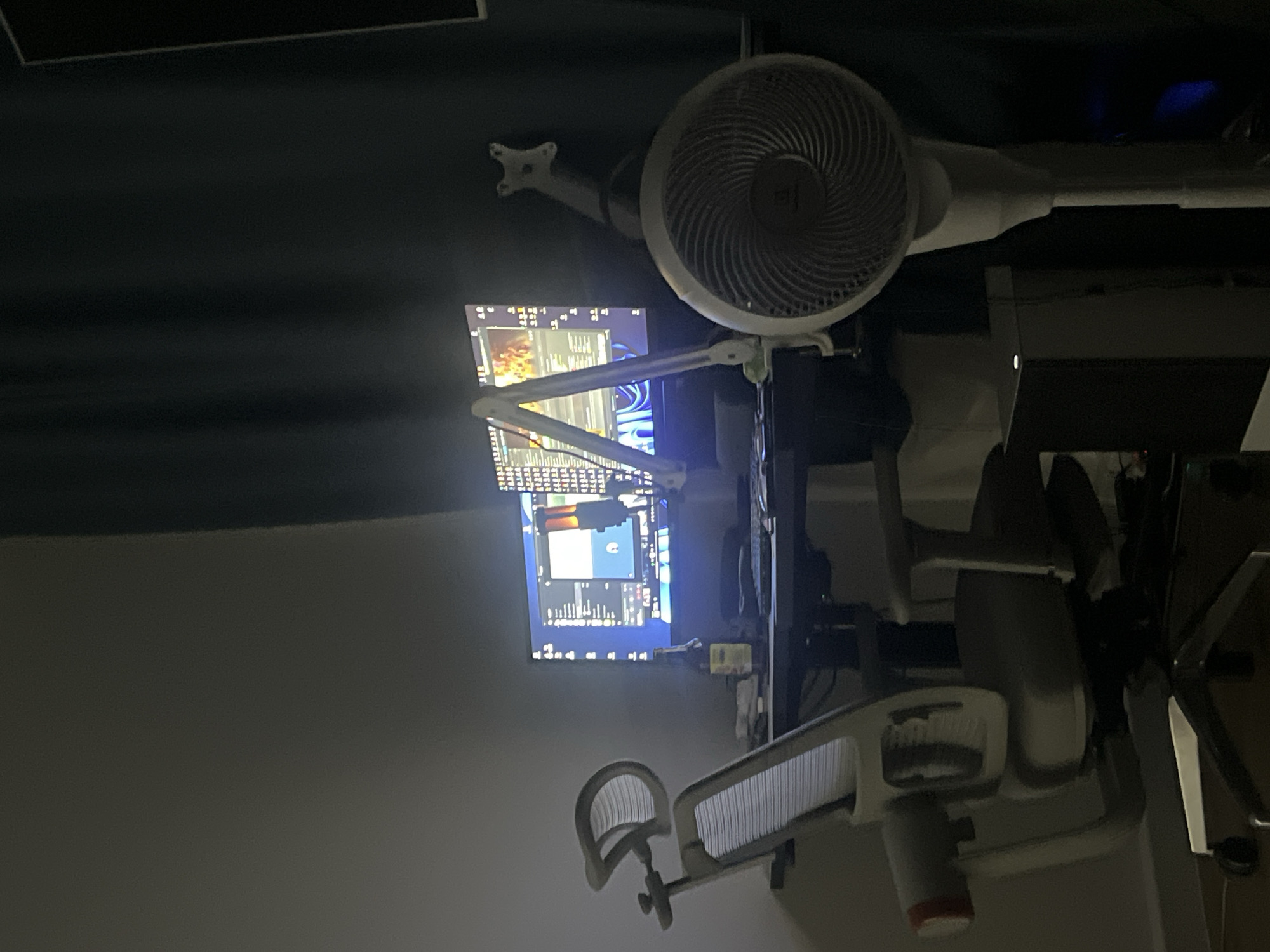 The image shows a home office setup in a dimly lit room. The main focus is a desk with a computer monitor, keyboard, and a microphone arm. An ergonomic office chair is positioned in front of the desk. To the right of the desk, a floor fan is visible. The background features a dark curtain and a light-colored wall. There are no people in the image, and no specific identifiable location markers are present.