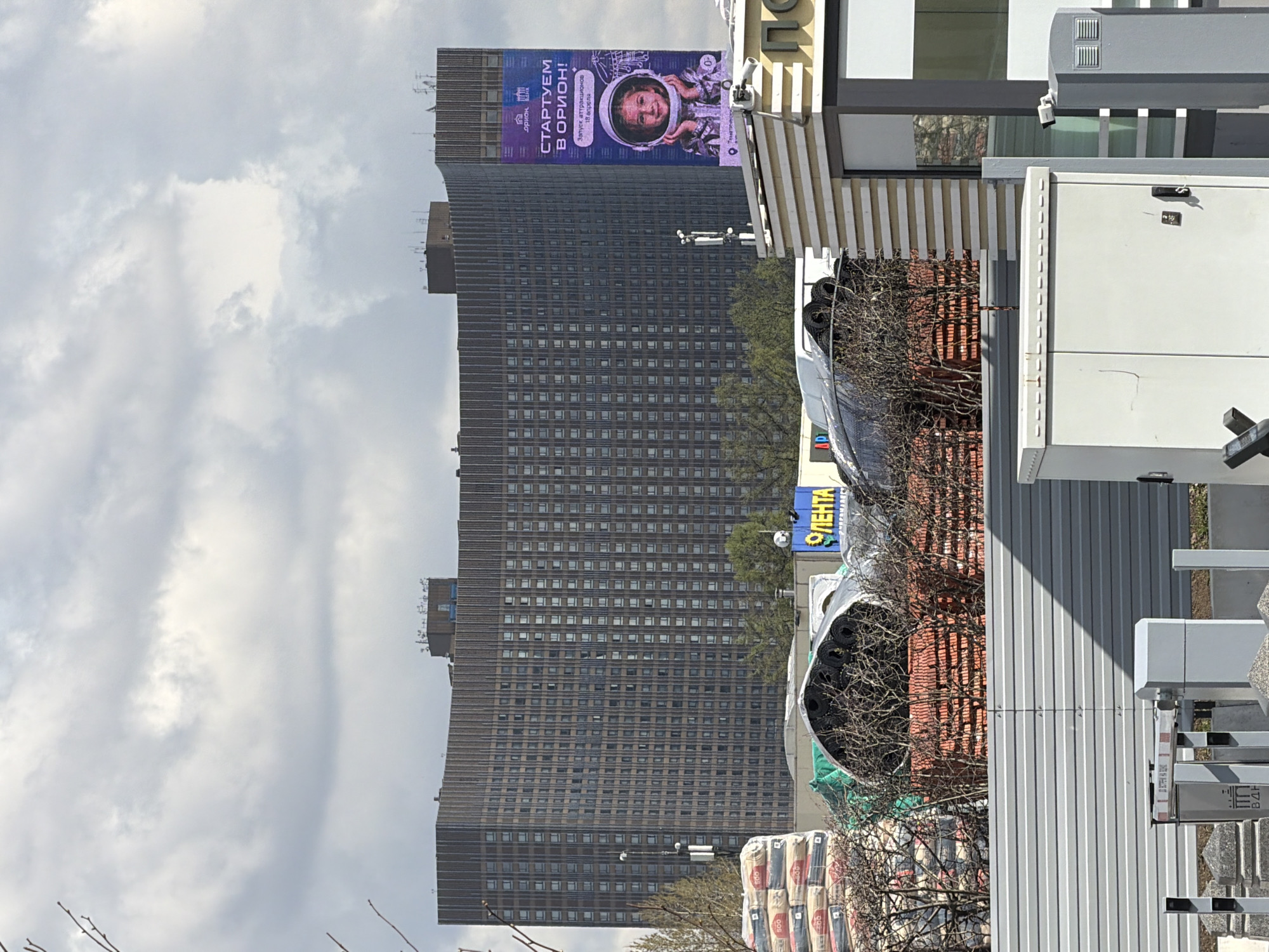 The image depicts an urban street scene in Moscow, Russia, during the daytime under an overcast sky. The focal point is a large, tall building dominating the background, possibly a hotel or office block, characterized by its numerous, uniformly arranged windows.
Attached to the upper part of this imposing building is a large, colorful billboard. The billboard features a cheerful young child wearing an astronaut's helmet, giving a thumbs-up gesture. Prominently displayed Cyrillic text on the billboard reads "СТАРТУЕМ В ОРИОН!" (STARTING IN ORION!), with smaller text below stating "Запуск аттракционов" (Launch of attractions) and "18 апреля" (April 18), indicating an event or attraction opening on that date. A logo with "ОРІОН" (Orion) is visible, likely identifying the venue.
In the foreground, several architectural and commercial elements are present. To the right, part of a building with a striped awning is visible, next to large, light-colored utility boxes or cabinets. Further into the foreground, there are rolls of what appear to be roofing or insulation materials covered with netting, and stacks of bagged goods, possibly construction materials. A yellow sign with the Cyrillic word "ЛЕНТА" (LENTA), a common Russian supermarket chain, is partially visible. Bare tree branches suggest either late winter or early spring. The overall impression is a bustling urban environment with ongoing commercial activity.