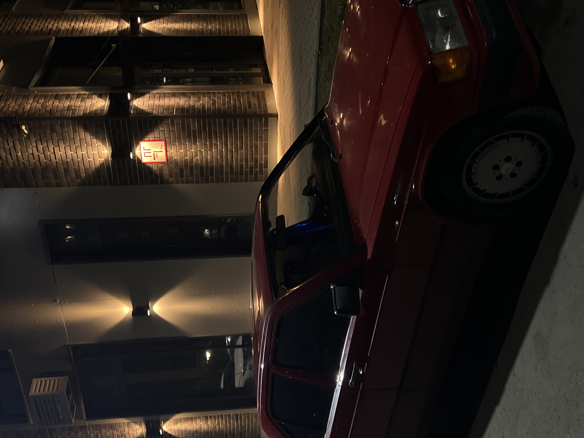 A red car is parked in front of a building at night. The building has a gray facade with horizontal rows of lights shining downwards. Part of the building has a brick facade. A sign is visible on the brick facade with what appears to be Cyrillic text. The car is parked on a paved surface. The image appears to be taken in a city, with architectural lighting suggesting a modern or urban environment. The city is Astana, Kazakhstan.