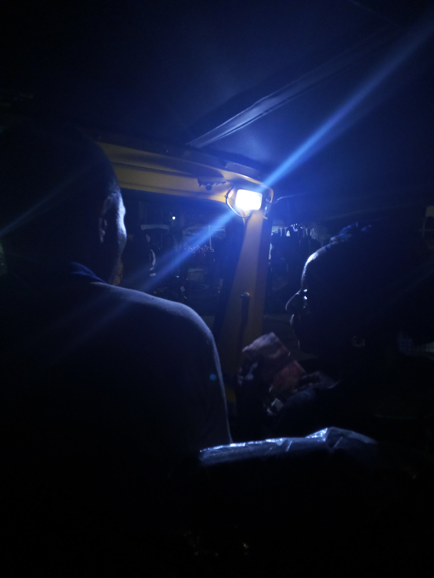 The image shows the interior of a vehicle at night, possibly a rickshaw or auto-rickshaw, as suggested by the presence of a bright headlight at the front. Two people are visible from behind, seated in the vehicle. The person on the left is facing away from the camera, with their head turned slightly to the right. The person on the right is facing the camera from a side profile, with their head turned to the left. Both individuals appear to be of African descent. The lighting is dim, with a strong blue light emanating from the vehicle's headlight, creating a beam that cuts across the dark interior and illuminates parts of the background. The background is indistinct but suggests a busy street environment with other lights and possibly some structures. The overall impression is one of movement and activity in a nighttime urban setting. The location in the city of Lokoja, Nigeria, cannot be determined from the visual information alone.
