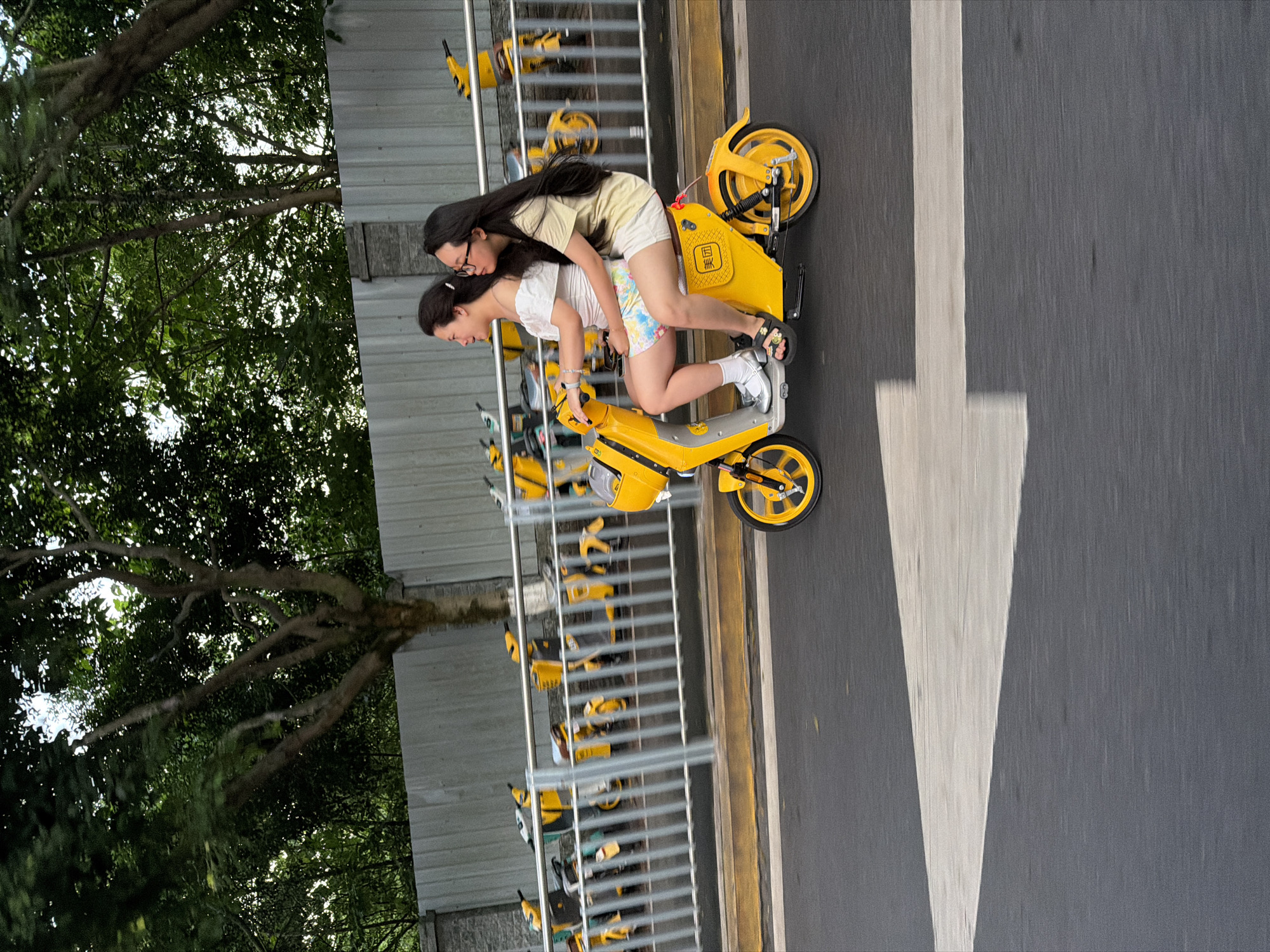 Two young women ride a yellow shared electric scooter on a road in the city of Chongqing High-tech Zone, China. The driver wears a white top and colorful shorts, and the passenger behind her wears a yellow shirt and white shorts. They are riding past a row of parked shared electric scooters behind a metal railing, next to a sidewalk with a tree. A large white painted arrow is visible on the gray asphalt road.