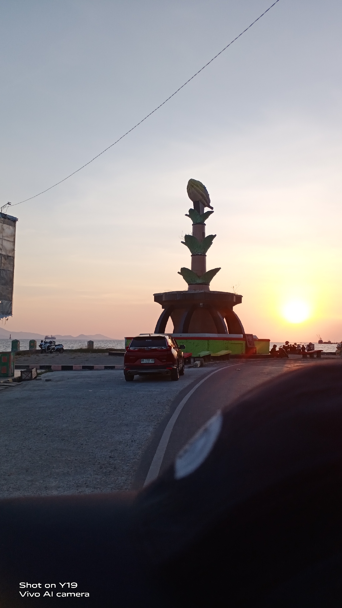 This image captures a sunset scene at a waterfront area, likely in Sea, Indonesia, given the provided text "Shot on Y19 Vivo AI camera" which suggests an Asian origin. A prominent monument, shaped like a stylized fruit or seed with green leaf-like accents, stands at the center-right of the frame. The monument is situated on a multi-tiered platform with red and green detailing.

In the foreground, a dark red SUV is parked on a paved road. The road curves towards the viewer on the right side, with a white lane marker visible. To the left of the monument, a body of water, presumably the sea, is visible with a few boats and a small vessel in the distance. Along the waterfront, there are several posts and what appear to be some rudimentary structures.

Several people are visible near the monument, some sitting and observing the sunset. The sky is a gradient of soft orange and yellow hues, indicating the sun is low on the horizon, casting a warm glow over the scene. A thin wire stretches diagonally across the upper portion of the sky. The overall atmosphere is calm and serene, typical of a coastal sunset.