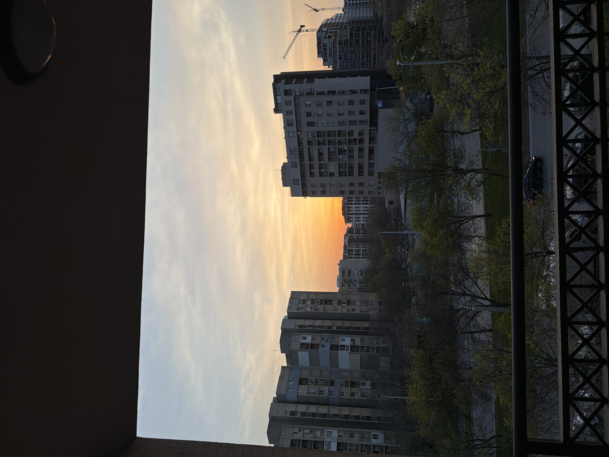 The image shows a view from a balcony in Plovdiv, Bulgaria, during a sunset. In the foreground is a dark balcony railing and a wall. The background contains several apartment buildings of varying heights and construction styles. A construction site with a crane is visible behind one of the buildings. A line of trees separates the buildings from a road with visible vehicles. The sky displays an orange and yellow gradient near the horizon, transitioning to a light blue higher up.