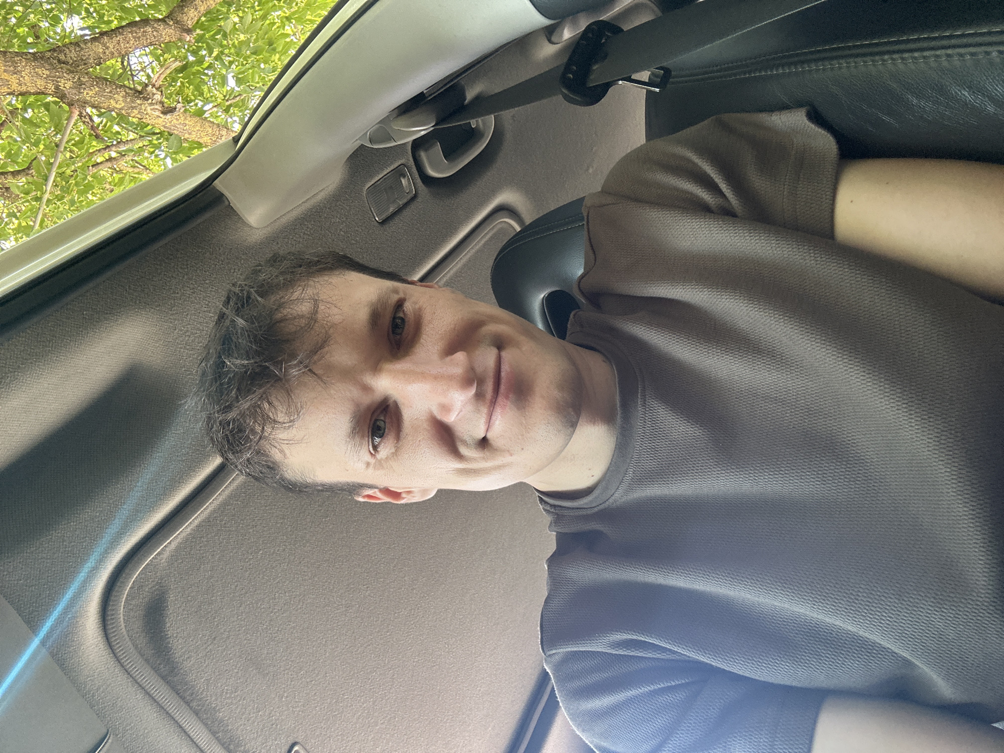 A selfie taken from inside a car shows a man looking at the camera. He is wearing a brown t-shirt. The interior of the car, including the ceiling and a seatbelt, is visible. Through the window, green leaves and tree branches are seen, suggesting an outdoor environment. There is no specific indication of the location being in the city of Makiv, Ukraine.