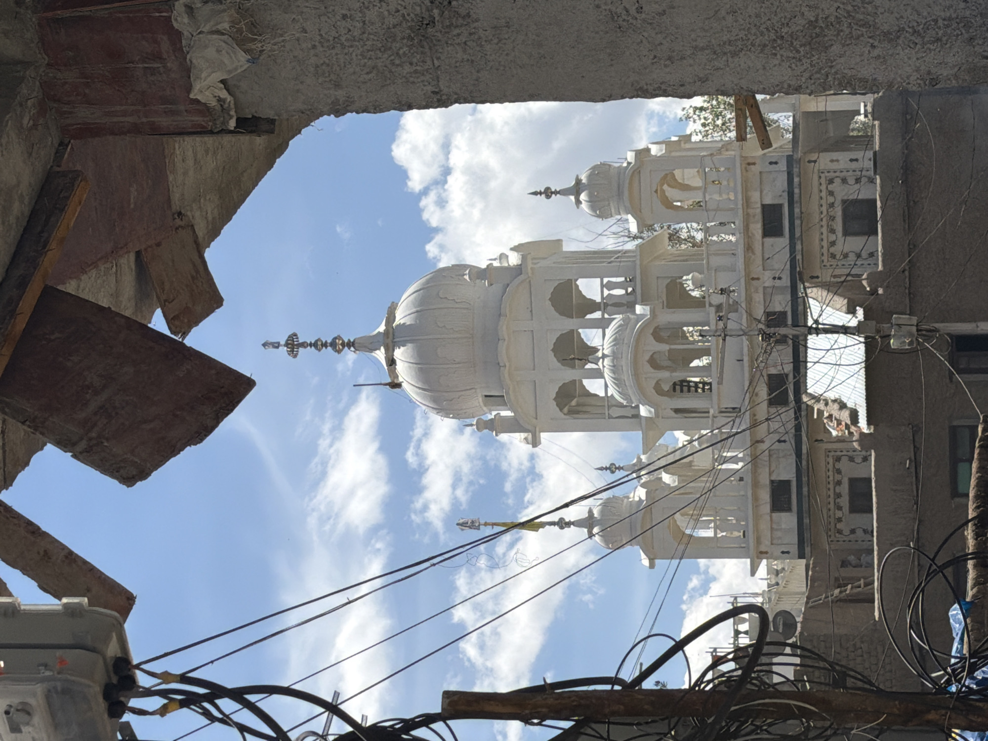 The image captures a portion of a white Sikh temple, likely a Gurdwara, in the city of Leh, India. The temple features traditional Sikh architectural elements, including domes, spires, and ornate detailing. It is framed by rough, unfinished concrete structures and a dense network of electrical wires and cables in the foreground, suggesting a juxtaposition of ancient spiritual architecture with the often haphazard urban development. The sky is blue with scattered white clouds, indicating fair weather. The temple appears to be a significant landmark, visible above other buildings in the city. The specific Gurdwara is likely the Gurdwara Pathar Sahib, a notable shrine in Leh.