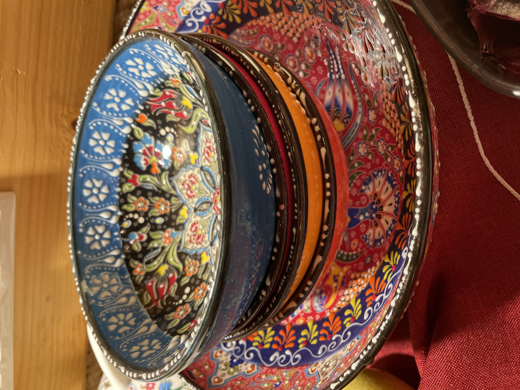 This is a close-up, high-angle shot of a stack of colorful, intricately decorated ceramic bowls. The bowls are nested inside one another, with only the rims and some of the interior patterns visible. The outermost bowl is a large, flat plate with a vibrant red background adorned with a complex floral and paisley design in blue, yellow, green, and white. The next bowl in the stack, partially visible, appears to have a dark blue base with a similar decorative pattern. The bowl directly in the foreground is a deep blue with a black interior, featuring a detailed floral pattern in white, red, green, yellow, and orange. The rims of all the bowls are edged with small, white decorative dots.
The setting appears to be indoors, with a wooden surface visible to the left and a textured red fabric to the right. The lighting is moderate, highlighting the glossy finish of the ceramics. There is no visible activity or event taking place; the image focuses solely on the arrangement and decoration of the bowls. The overall impression is one of artistry and craftsmanship, with a rich display of traditional patterns and vibrant colors. The image does not contain any visible text.