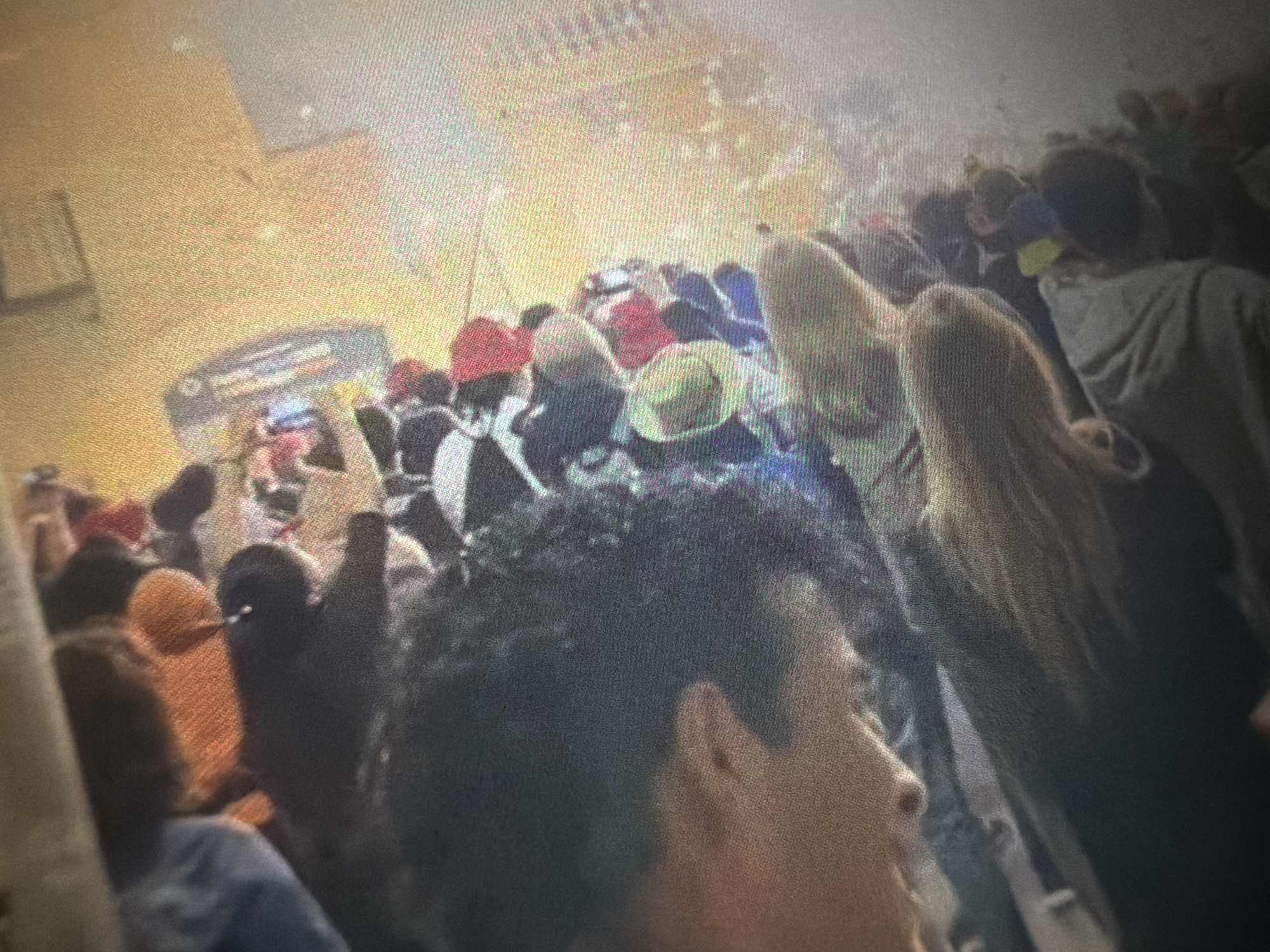 A grainy, slightly blurry image captures a dense crowd of people gathered in what appears to be a large indoor public space. The perspective is from slightly above and behind the crowd, looking towards a general direction where their attention is focused.
**Subjects:** Numerous individuals, both men and women, are visible, predominantly from their backs and heads. Hair colors range from dark curly to lighter blonde.
**Objects:** The crowd is dressed in casual attire, including hoodies, t-shirts, and several hats, notably red caps and at least one straw-colored hat. In the background, a large, blurry sign is visible.
**Scene/Setting:** The setting is an interior space, characterized by bright, artificial lighting. The walls and ceiling appear light-colored and somewhat indistinct. The density of the people suggests a significant event or gathering.
**Activity or Event Taking Place:** The people are standing close together, indicating a crowded environment such as a concert, convention, or public event. Their collective gaze towards a common point implies they are observing or participating in an activity. Some individuals appear to have their hands slightly raised, possibly in a gesture of applause, waving, or engagement.
**Notable Details:** The image quality suggests it might be a photograph of a screen, contributing to the pixelation and slight blurriness. The lighting is consistent and bright, with no obvious shadows indicating a specific time of day beyond being well-lit indoors. The location context of Sant Martí Sarroca, Spain, suggests this could be at a local public venue.
**Visible Text:** A prominent sign in the upper left background clearly displays the text "FOOD COURT". Above this, partially obscured but discernible, is the word "PALAU", likely part of a larger name like "Palau de Congressos" (Convention Palace) or similar, further confirming the indoor public event setting.