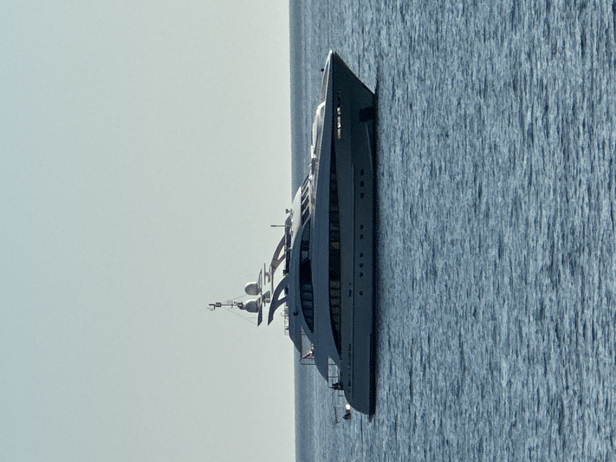 A large, light-colored luxury yacht is anchored in calm, blue water. The yacht has multiple decks and sleek lines, with numerous windows visible along its hull and superstructure. At the top of the yacht, there are radar dishes and antennas. The sky above is a pale, cloudless blue. The yacht is positioned in the foreground, dominating the frame, with the sea and sky occupying the rest of the image. No people are visible on the yacht or in the surrounding water. The scene appears to be on a clear, bright day, likely in a coastal area. The image does not contain enough information to determine its specific location within Budva, Montenegro, beyond it being a maritime setting.