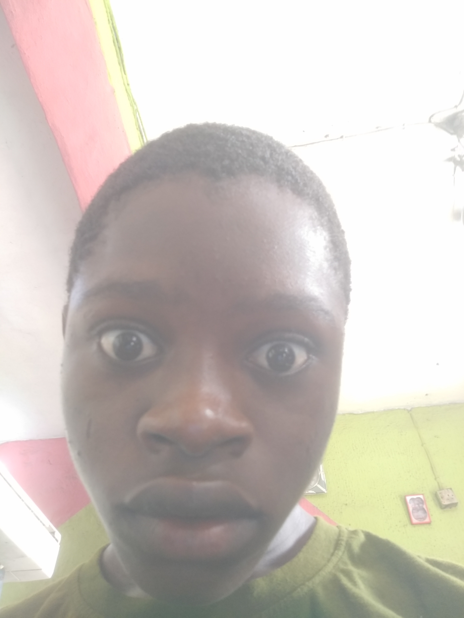 A close-up selfie of a young person with dark skin and short, dark hair, facing the camera with wide eyes and a slightly open mouth. The person is wearing a greenish-brown shirt. The background shows a wall painted with a horizontal stripe of pink and yellow near the ceiling, and a green lower portion. On the green wall, there's a framed picture, a rectangular object, and an electrical outlet. The photo appears to be taken indoors. The location in Shangisha, Nigeria, is not explicitly confirmed but inferred based on the context.
