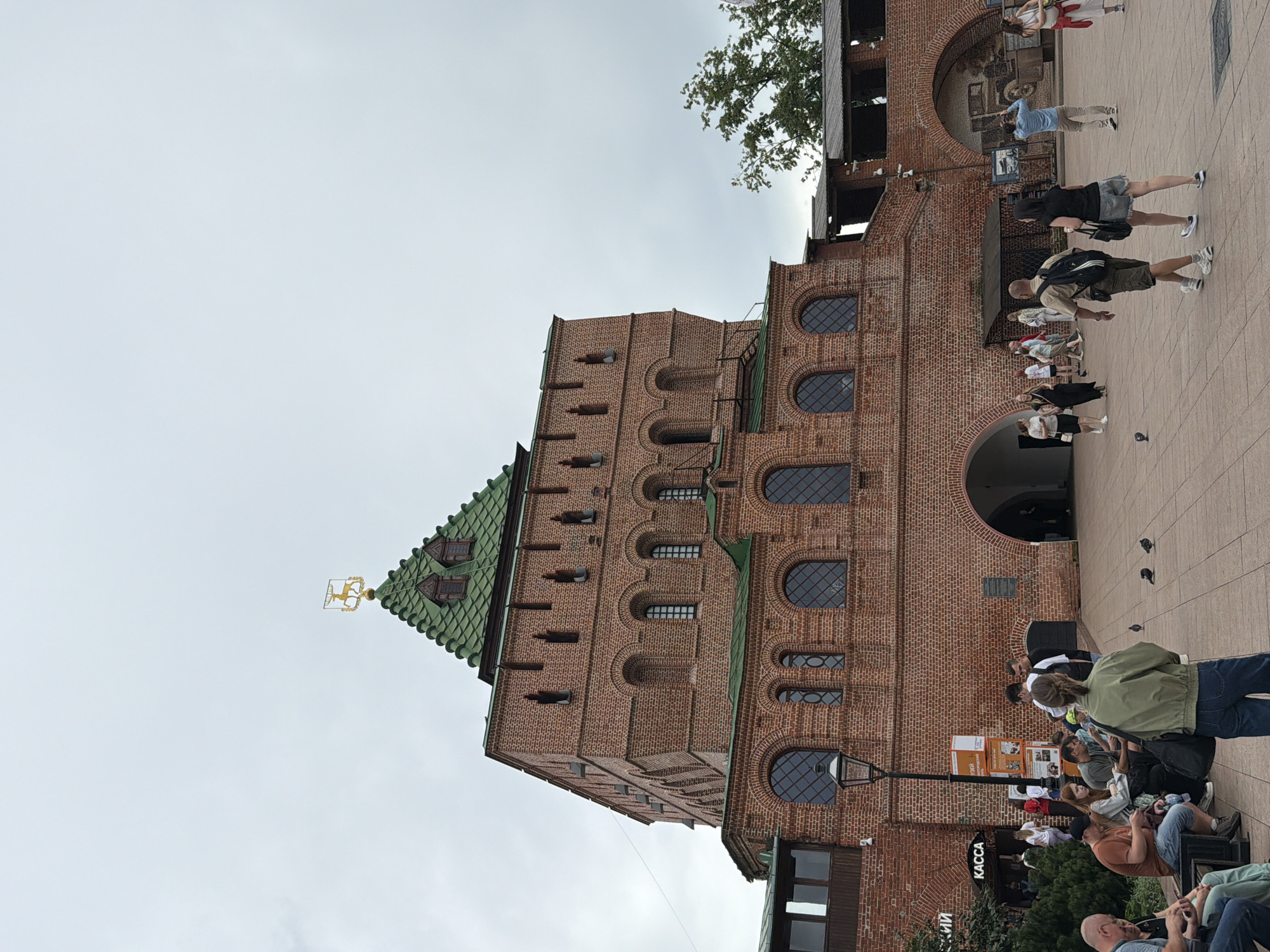 The image depicts a view of the Nizhny Novgorod Kremlin, specifically the St. George's Tower. The tower is constructed of red brick with a green tiled roof, topped by a golden weather vane shaped like a horse. People are visible in the foreground, some sitting on benches, others walking, and a few are standing in groups. Some are looking at their phones, while others appear to be conversing. There is a "KACCA" (cashier) sign visible on a building adjacent to the tower. The ground in front of the tower is paved with square tiles. The sky is overcast. The overall scene suggests a public gathering space within a historical site.