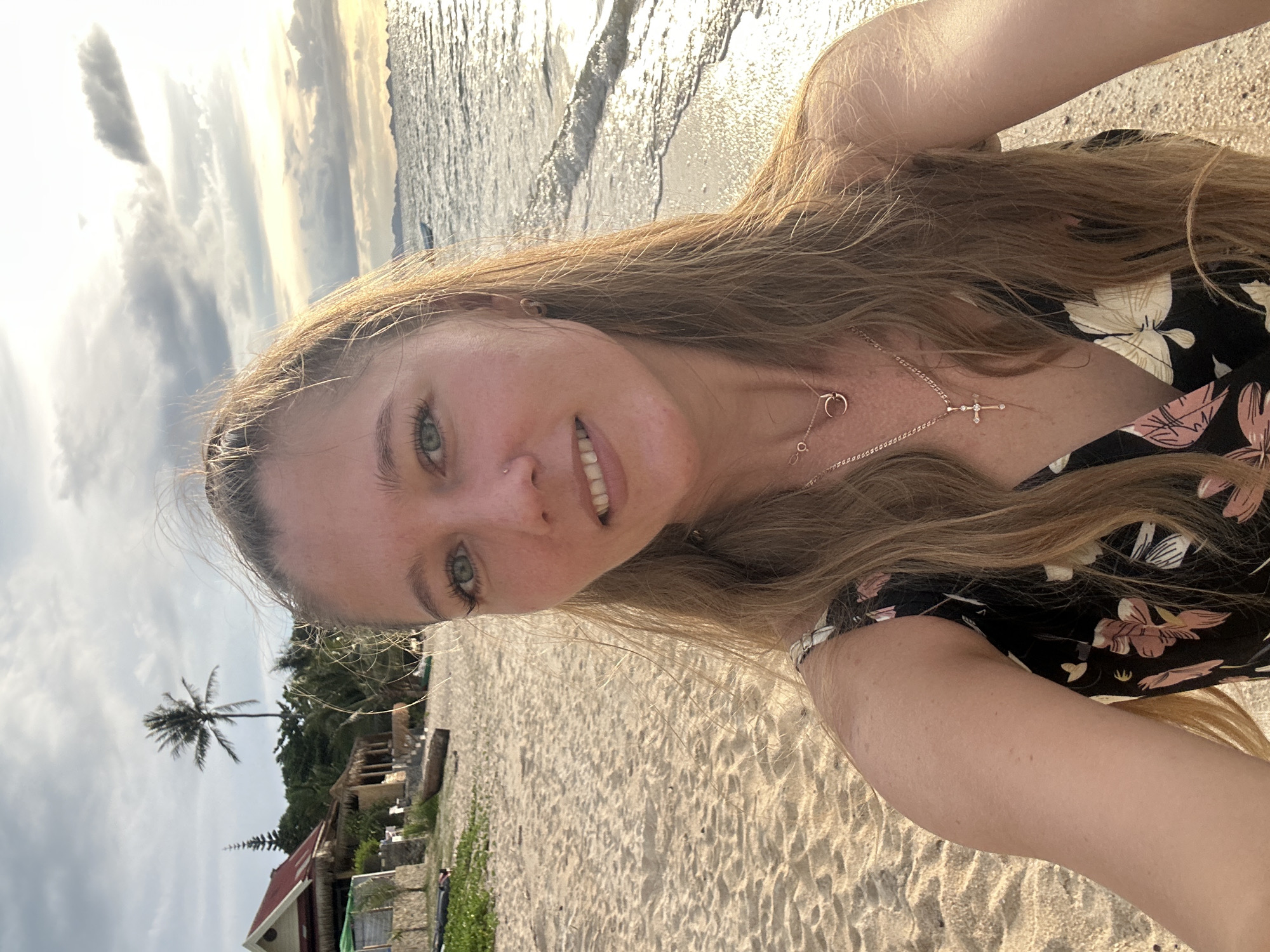 A woman is taking a selfie on a beach in Baan Thong Po, Thailand. The woman has long blonde hair and is wearing a black dress with a floral pattern. She is smiling at the camera. The beach is sandy and there are palm trees and some buildings in the background. The ocean is to the right of the woman, and there are waves gently breaking on the shore. The sky is cloudy with the sun setting, casting a warm glow over the scene.