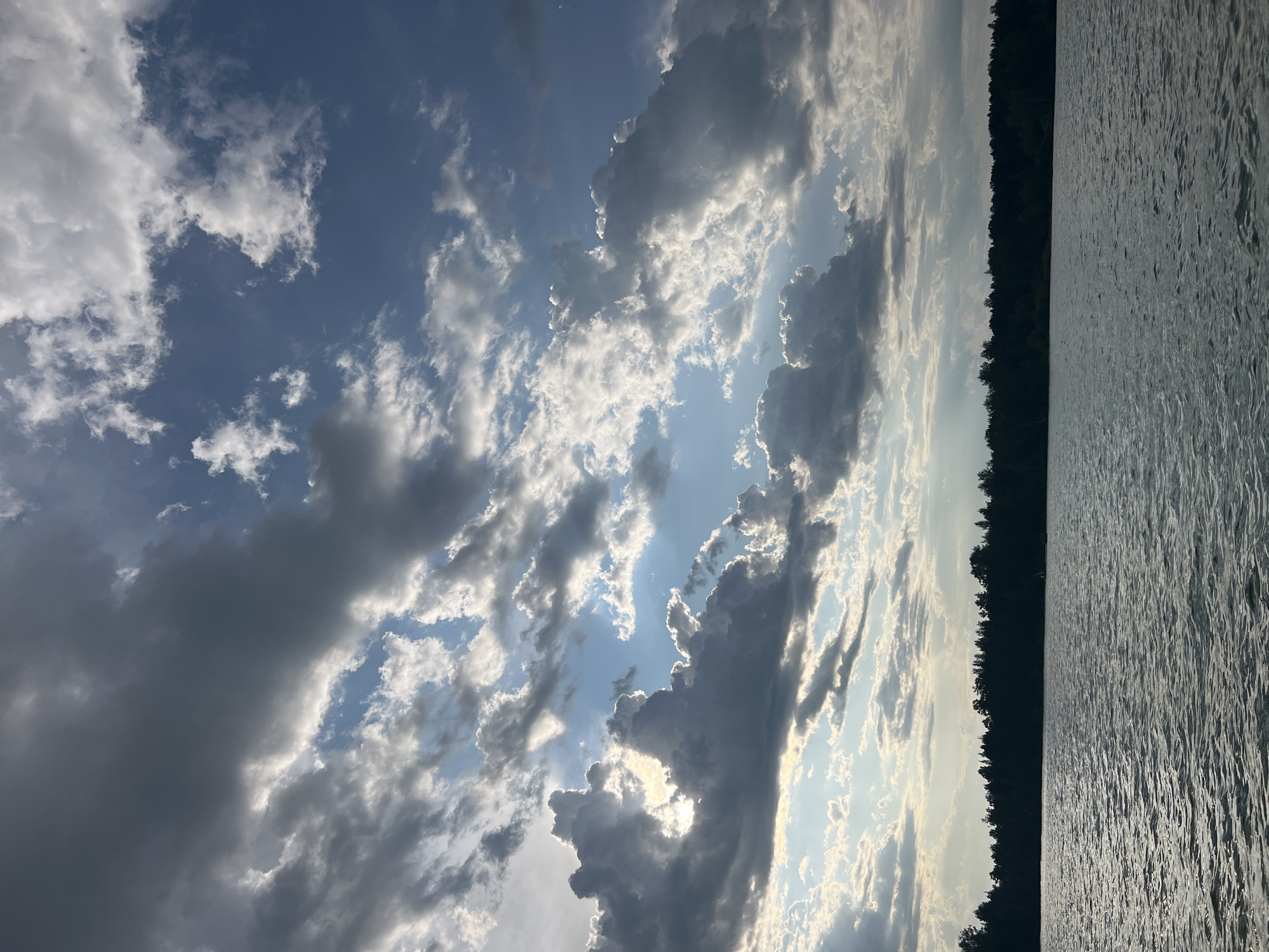 A dynamic sky with a mix of dark, brooding clouds and bright, sunlit patches hovers over a serene body of water. The sun's rays pierce through gaps in the clouds, creating dramatic shafts of light that illuminate the sky. Below the sky, a dark, silhouetted treeline stretches across the horizon, dividing the sky from the rippling surface of the water. The water itself is choppy, reflecting the changing light and dark patterns of the sky. The scene appears to be a natural landscape, possibly a lake or a wide river, bordered by dense forest. There are no people or man-made structures visible in the image, suggesting a remote or natural setting. The overall impression is one of dramatic natural beauty, with the interplay of light and shadow creating a sense of depth and atmosphere.