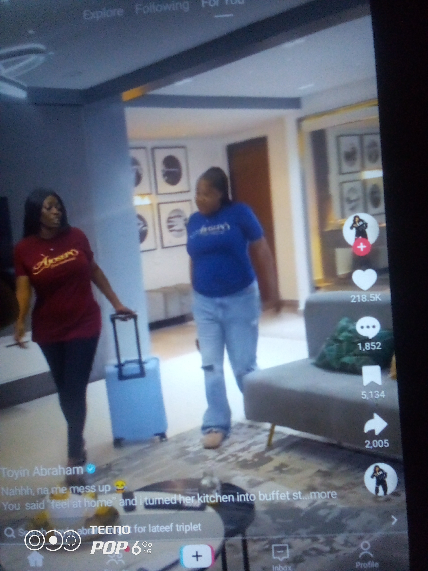 Two women are seen in a well-lit interior space, possibly a modern apartment or hotel lobby. The woman on the left wears a maroon t-shirt with yellow lettering and dark pants, pulling a light blue suitcase with black trim. The woman on the right is dressed in a blue t-shirt and light-wash distressed jeans. The background features framed artwork on a wall and a grey sofa with a plant. The scene appears to be captured on a mobile device, as indicated by the on-screen interface elements, including icons for likes, comments, shares, and profile views, as well as text overlays identifying "Toyin Abraham" and other comments. The city of Howayi, Nigeria, is not discernible from the visual information in the image.