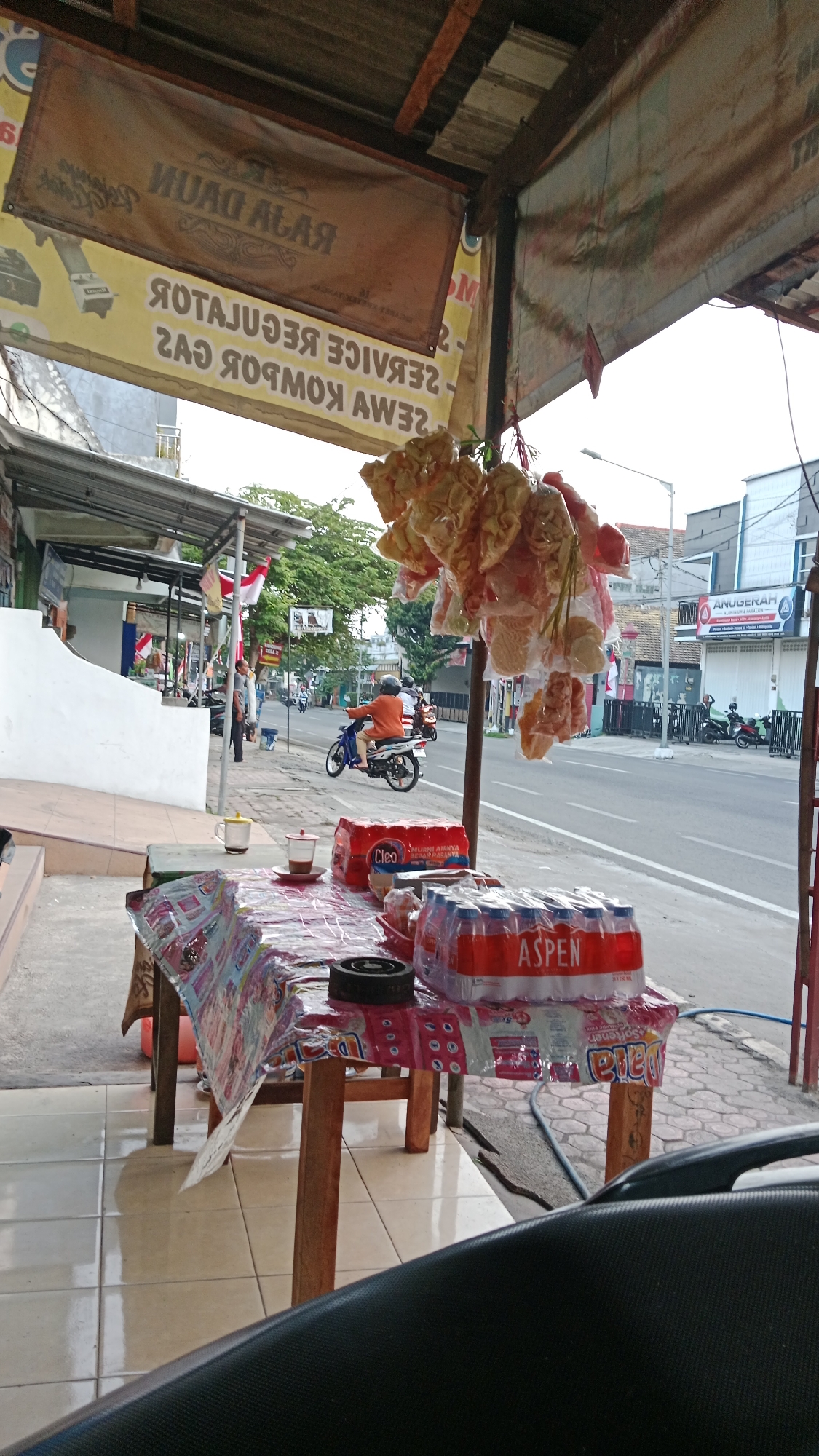The image captures a street-level view of a roadside stall in Blitar, Indonesia. In the foreground, a wooden table is covered with a colorful, patterned plastic tablecloth. On the table, there are two cups, one with coffee and a saucer, and a stack of bottled water, branded "ASPEN". A small, round object, possibly an ashtray, is also on the table. Hanging above the stall are several plastic bags filled with snacks or crackers, tied together with string.
In the mid-ground, a person wearing an orange shirt and a helmet is riding a blue and white motorcycle on the street. The street is paved and appears to be a busy thoroughfare with other vehicles and buildings lining its sides. The buildings have awnings and signage, with one prominent sign in the background reading "ANUGERAH".
Further in the background, trees and more buildings are visible. The overall environment suggests a typical urban street scene in Indonesia, likely a commercial area with shops and food vendors. The presence of Indonesian flags indicates a possible celebration or national event. The perspective suggests the photo was taken from a seated position, possibly on a motorcycle or scooter.