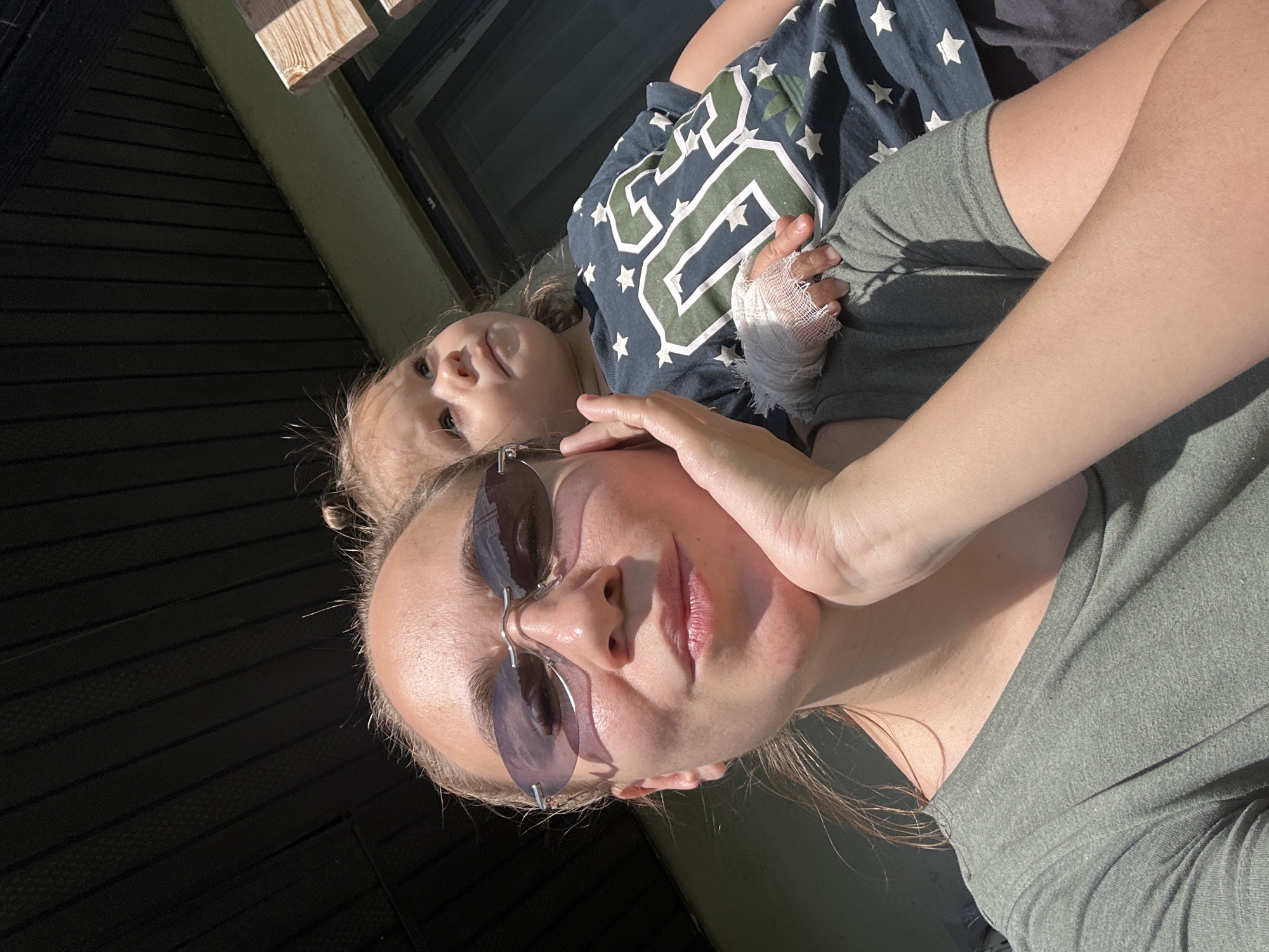 The image, captured outdoors in Kharnavy, Russia, depicts an adult female and a young child under direct sunlight. The adult, positioned in the lower-left to mid-frame, wears rimless sunglasses with purple-tinted lenses and an olive green short-sleeved t-shirt. Her right hand is placed under her chin, appearing to support her head. The child, situated to the right and slightly behind the adult, possesses light-colored hair and wears a dark blue t-shirt patterned with white stars and a prominent green "03" graphic. The child's left hand is bandaged with white gauze and rests on the adult's upper arm. The adult faces the camera, while the child gazes upwards and to the right. The background features a dark, planked overhead structure, a section of light-colored wooden railing or beam, and a dark window frame, indicative of a residential outdoor environment.