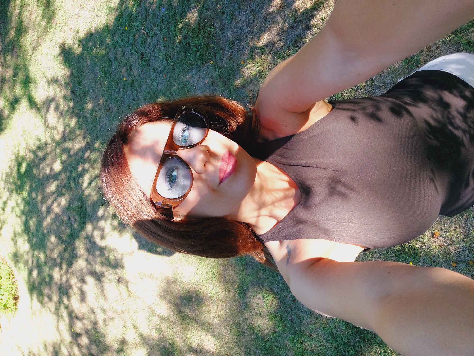 A woman with auburn hair and blue eyes, wearing large brown-rimmed sunglasses and a dark brown sleeveless top, takes a selfie outdoors. She is standing in a grassy area with dappled sunlight filtering through trees, creating shadows on the ground and on her. Her lips are slightly parted and painted with pink lipstick. A tattoo is visible on her left shoulder. She is wearing white pants.