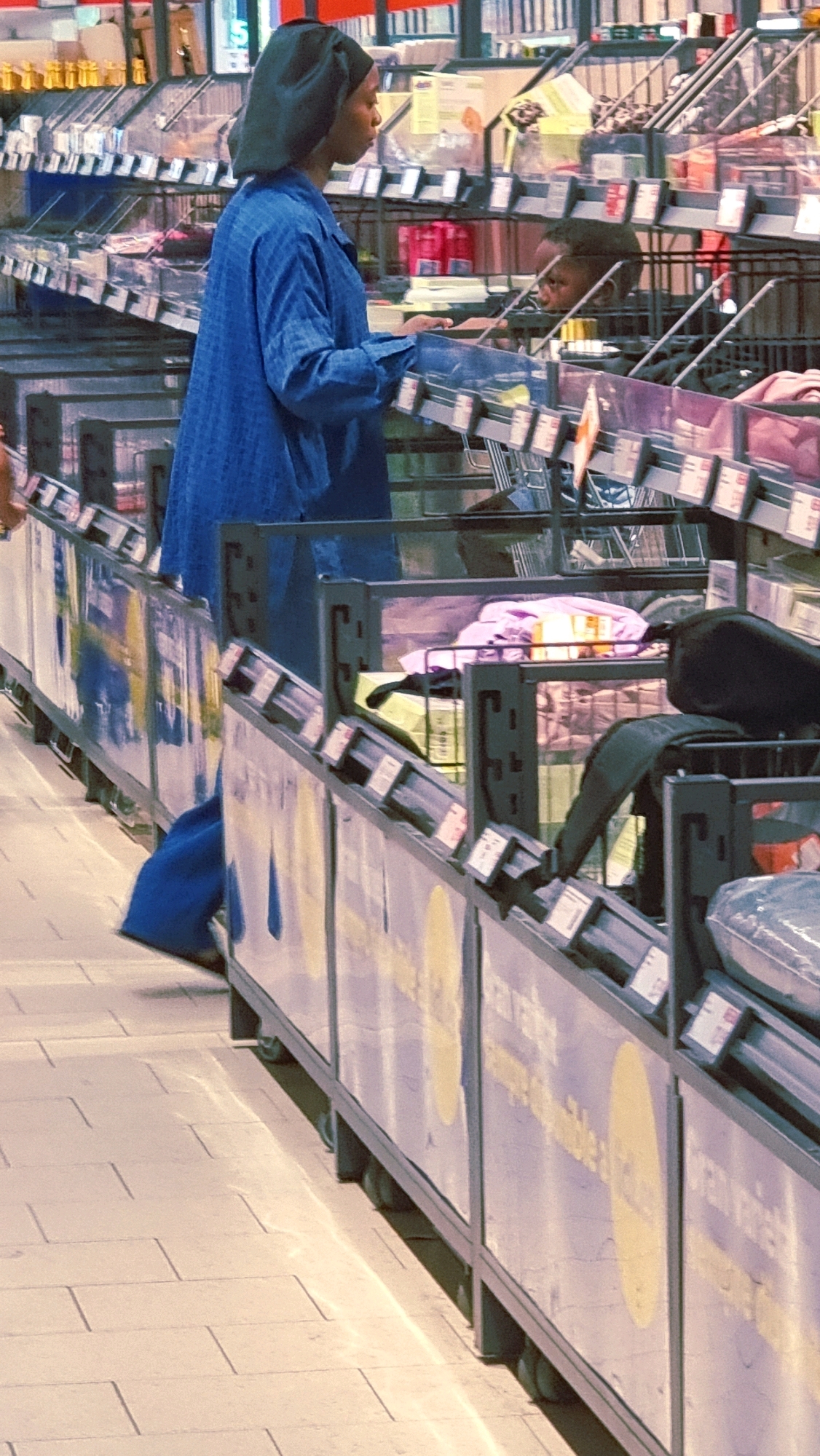 The image depicts an indoor scene, likely a supermarket or a market stall, with rows of products displayed on shelves. In the foreground, a person wearing a dark head covering and a blue patterned long-sleeved garment is visible from the waist down, pushing what appears to be a shopping cart or display unit. The person's left leg is extended, and they are holding the unit with both hands. The background shows more shelves stocked with various items, some in transparent packaging. The flooring is tiled, and sunlight appears to be reflecting off it, creating streaks of light. There is another person partially visible in the background, their face turned to the side. The overall impression is a commercial setting with goods for sale. The city of Olot, Spain, is not identifiable from the visual cues in the image.