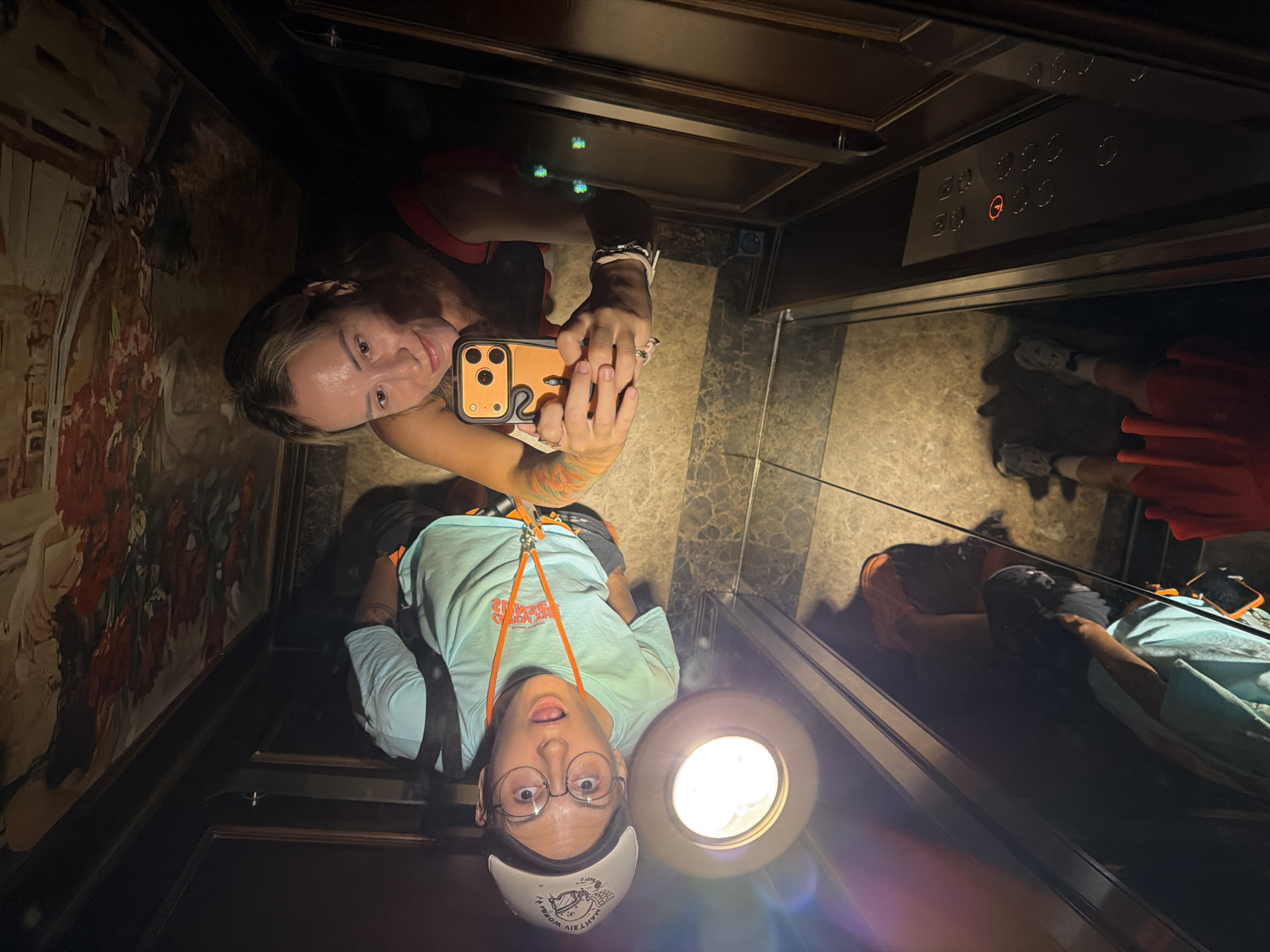 In an elevator with mirrored walls and a decorative ceiling, two individuals are captured in a selfie. The person on the left wears a light blue t-shirt, glasses, and a white head covering with a crescent moon design. They are looking upwards with their mouth open. The person on the right, wearing a red and black top, holds an orange smartphone and is taking the photo. Their arms, adorned with tattoos, are visible. The elevator floor is tiled with a marbled pattern. The reflection in the mirror shows another individual, partially obscured, wearing a light blue shirt and dark pants, and the lower legs of two people wearing sneakers and red shorts. The elevator panel with buttons is visible on the right side, with a red indicator light illuminated on button labeled "1". The image appears to be taken in the city of Thuận Hóa Ward, Vietnam, based on the provided context.