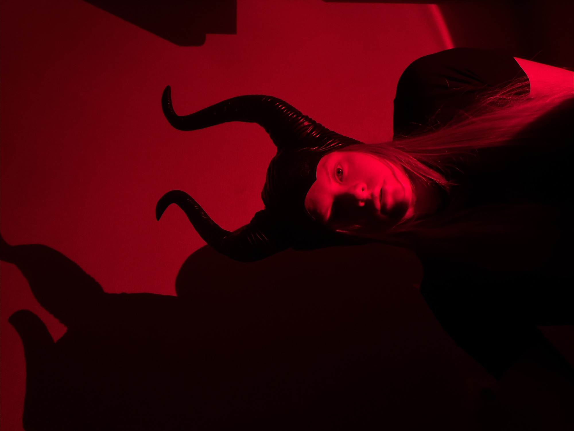 A dramatic, high-angle portrait features a young person, likely a woman, wearing a dark, textured horned headpiece. She is lying down or leaning back, looking directly at the viewer with a neutral and serious expression. Her face and visible light-colored hair are brightly illuminated by a strong, artificial red light, creating a stark contrast with the dark costume and the deep shadows it casts. The horns are prominent, appearing black with visible ridges.
The entire scene is bathed in this intense red light, which serves as both the primary illumination and the background color. The background is simple, appearing to be a solid wall or surface, entirely drenched in red. A large, dark shadow of the horned figure is cast on the lower left portion of the frame. There are also angular shadows in the upper left, suggesting other objects or structures in the room.
The image captures a posed moment, likely a costume or artistic shoot, emphasizing a mysterious and theatrical atmosphere through its dramatic lighting and subject's attire. There is no visible text in the image. The location context for this photograph is Izhevsk, Russia.