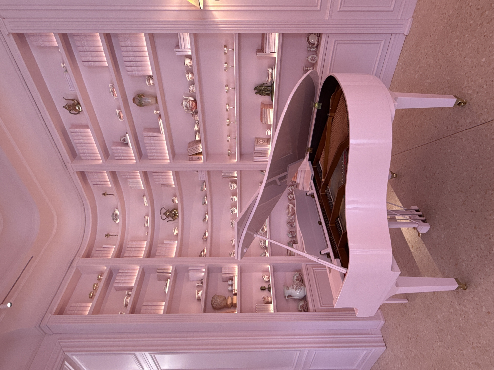 The image depicts a pink grand piano positioned in a room with curved, built-in bookshelves filled with decorative items and books. The room has a soft, warm lighting, casting a pink hue over the entire scene. The floor is a speckled terrazzo in shades of pink and beige. The overall aesthetic is elegant and whimsical. The image does not contain any information about the city of Kyiv, Ukraine.