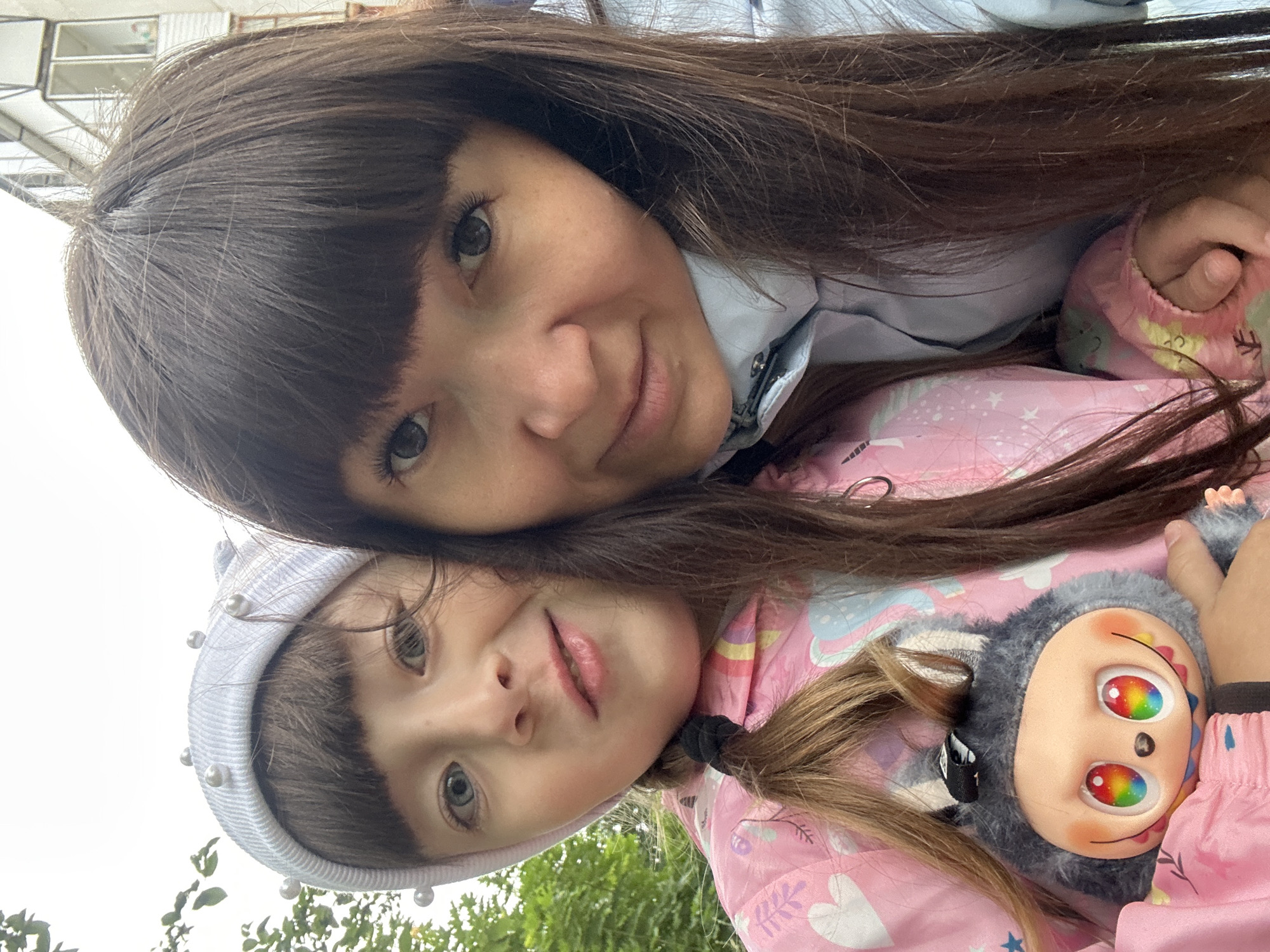 In this close-up selfie taken outdoors, two females are prominently featured. The individual on the left, a child, wears a white beanie adorned with pearls and a pink jacket with cartoon-like illustrations. Her hair is braided, and she holds a gray plush toy with a distinctive face featuring rainbow-colored eyes. Beside her, the individual on the right, an adult, has long, dark brown hair and wears a light blue jacket. They are positioned in a way that suggests they are together, with the child slightly in front of the adult. The background reveals a building with balconies, characteristic of urban environments, and some greenery, hinting at a residential area in Chaikovsky, Russia. The lighting suggests an overcast or diffused daylight condition.