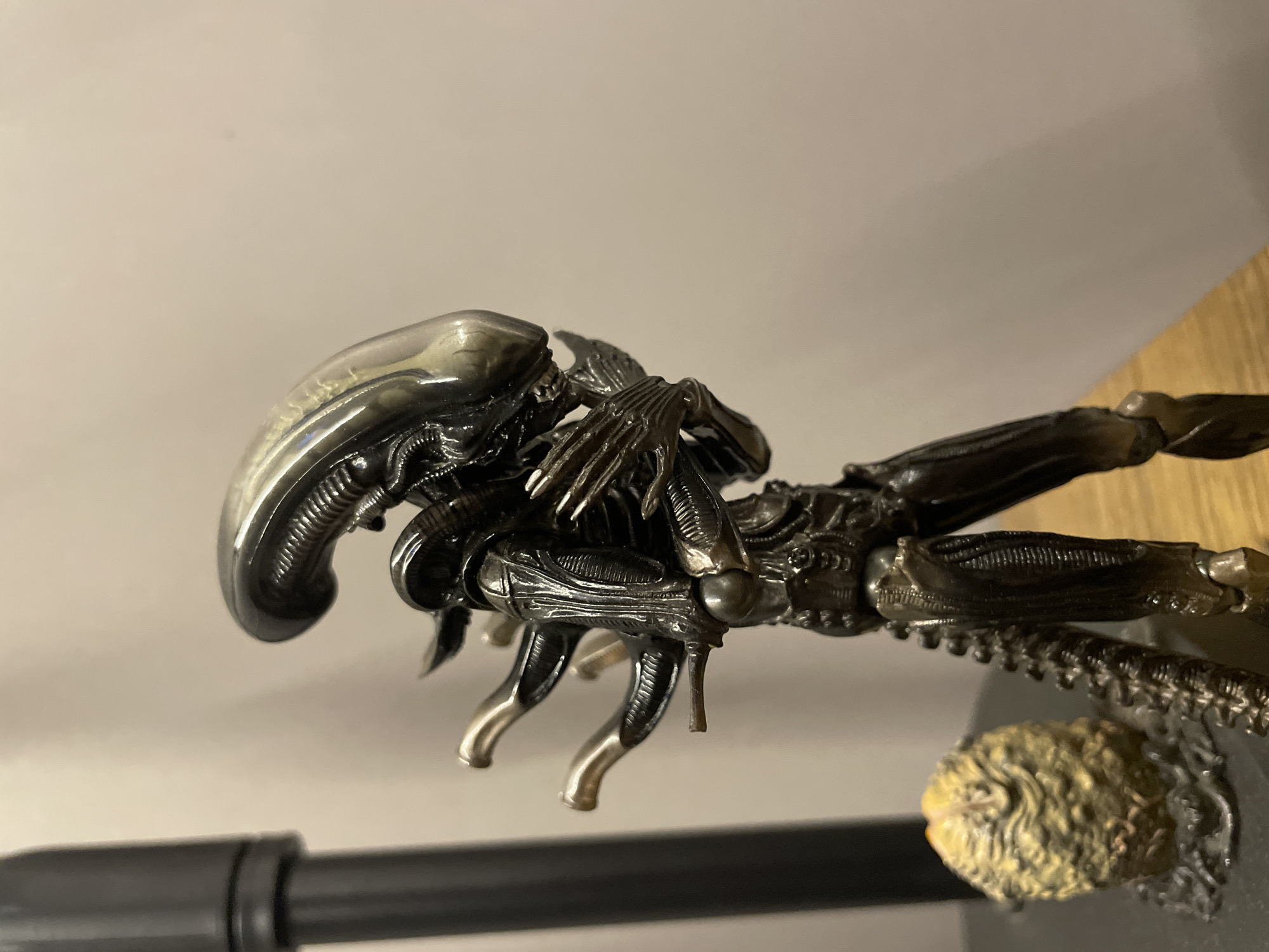 The image shows a Xenomorph action figure, likely from the "Alien" film franchise. The figure is primarily black and silver with a smooth, elongated head. It is posed in a crouching position with its limbs extended. The figure is on a dark surface with a textured, yellowish object visible behind it. In the background, there is a light-colored wall and a wooden surface. No location is discernible from the image.