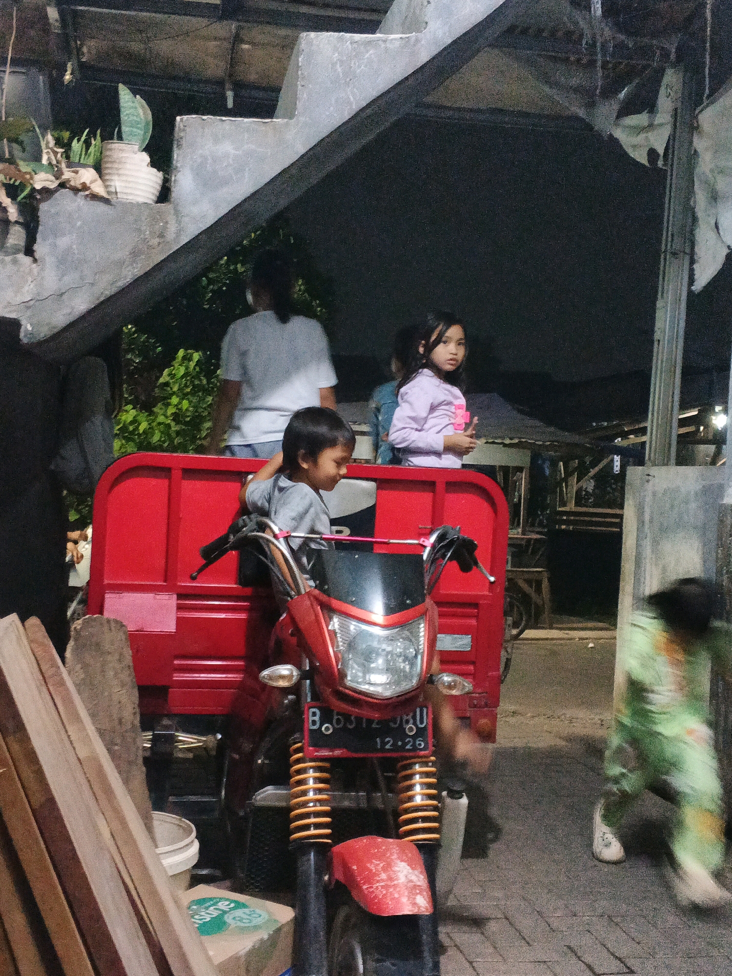 In Kadu, Indonesia, at night, two young children are seated in the back of a red three-wheeled motorcycle-like vehicle. The girl, wearing a light purple long-sleeved shirt, looks towards the camera, while the boy, in a grey t-shirt, leans his elbow on the vehicle's side and smiles to his left. Behind them, a person wearing a white t-shirt stands in the shadows. To the right, another child in a green outfit is in motion, partially visible, walking on a paved surface. The vehicle has a black and red front with visible license plate "B.B312 JKU" and "12.26". The background consists of a concrete staircase structure, some greenery, and what appears to be a stall or structure with tarpaulin roofing. Piles of lumber are visible in the foreground to the left.