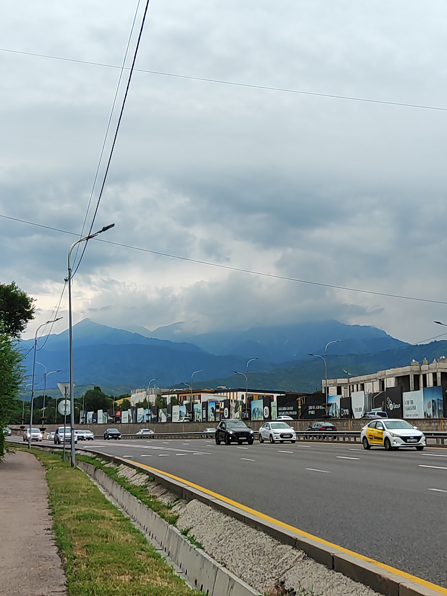 The image depicts a cloudy day in Almaty, Kazakhstan, with a highway in the foreground and mountains in the background. On the highway, several cars are driving. One of the cars is a yellow taxi. In the background, construction can be seen, along with a row of tall trees. The sky is filled with heavy, grey clouds, suggesting a potential for rain. The mountains in the distance are a hazy blue, possibly due to atmospheric conditions or distance. Power lines are visible across the sky, indicating the presence of urban infrastructure. The roadside is lined with grass and concrete barriers. The overall scene suggests a typical urban environment on a somewhat overcast day.