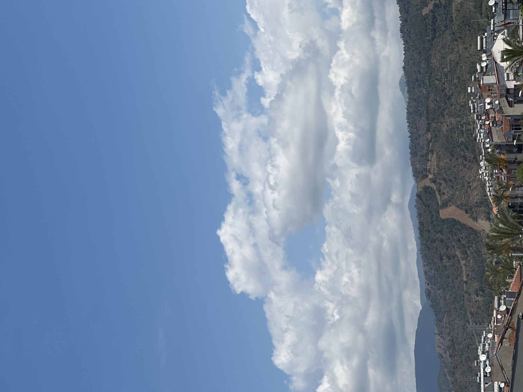 The image depicts a view in Fethiye, Turkey, characterized by a combination of natural and urban elements. The sky is predominantly blue, with a large mass of white clouds filling the upper right and center of the frame. These clouds have a dense, textured appearance. Adjacent to the clouds is a steep mountain range covered with trees and vegetation. At the base of the mountain, there is a residential area with closely packed buildings. Each building appears to have numerous satellite dishes and solar water heaters on the roofs. Palm trees are scattered along the edge of the rooftops.