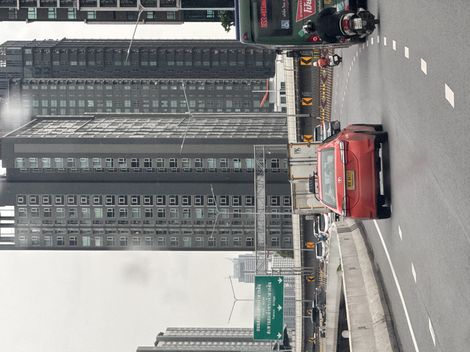 This is an eye-level, medium shot of a highway scene in Bangkok, Thailand, under an overcast sky. The focus is on a red sedan with a yellow license plate and the word "TAXI" on its roof, driving in the right lane. Behind the sedan, a motorcycle with a rider in a black jacket and helmet is in the adjacent lane. Further ahead, a green bus with visible text in Thai script is in the far right lane. The environment features several tall, modern apartment buildings in the background, characterized by their grey facades and numerous windows. A green directional sign with Thai and English text, indicating "Taksian Road" and "Taksian Bridge," is visible on the left. The road surface is asphalt with white lane markings. Traffic appears to be moderate, with other vehicles present in the background. The overall impression is a typical urban highway commute in Bangkok on a cloudy day.