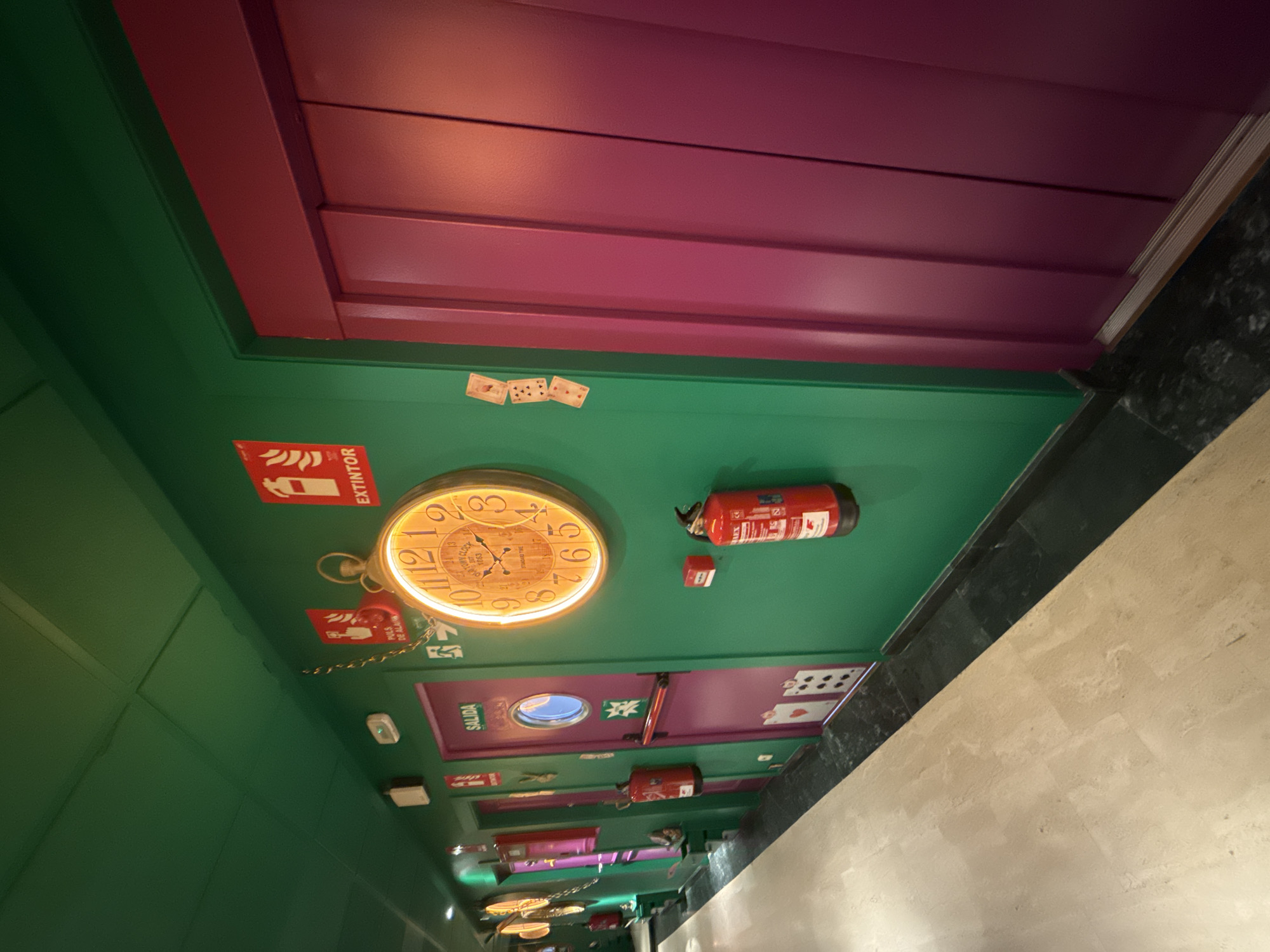The image captures a skewed perspective of a brightly colored ceiling in what appears to be a themed establishment, possibly in Madrid, Spain, given the Spanish signage. The ceiling is painted in vivid green and purple, with elements designed to create a whimsical or fantastical atmosphere. Prominent features include a large, illuminated decorative clock, a fire extinguisher, and various signage, including one indicating "SALIDA" (exit) and another indicating "EXTINTOR" (fire extinguisher). The ceiling is adorned with playing card designs, adding to the themed environment. The presence of emergency equipment suggests the location is a public space, such as a restaurant, bar, or themed attraction.