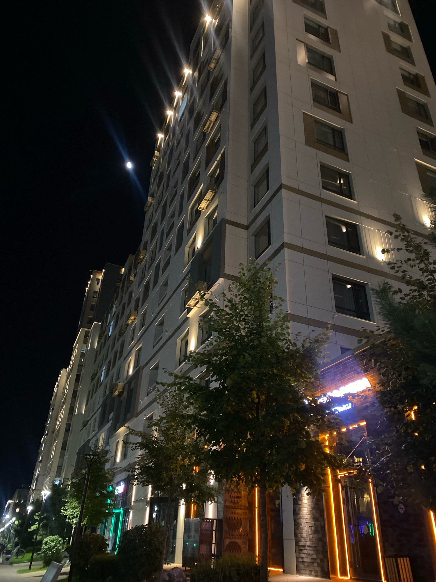 This is a low-angle, night-time photograph of a modern, multi-story building in Astana, Kazakhstan. The building, likely residential or commercial, features a light-colored facade with prominent horizontal bands and numerous windows. The top floors are illuminated by warm spotlights, casting rays into the dark sky where a full moon is visible. The lower floors of the building feature shops or businesses with brightly lit signage and illuminated decorative elements. Trees with green foliage are positioned in front of the lower sections of the building, and their branches partially obscure the view of the storefronts. The street in front of the building is also illuminated by streetlights, and a sidewalk is visible. The overall impression is one of a well-lit, contemporary urban environment at night.
