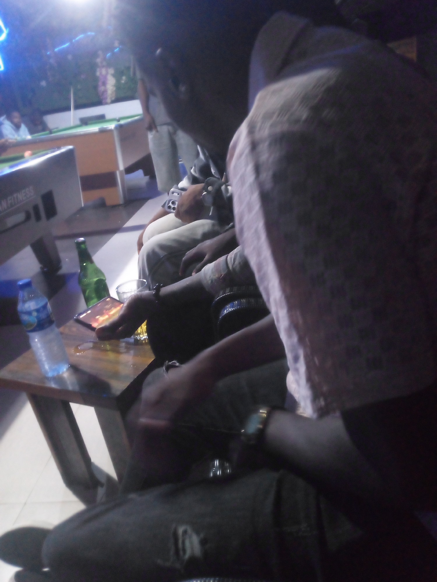 In a dimly lit indoor establishment, possibly a bar or lounge in Jos, Nigeria, a group of individuals are gathered around a table. The focus is on a person in the foreground, whose arm and shoulder are prominent. They are wearing a light-colored, textured garment. On the table, a plastic water bottle, a green bottle that appears to be a beverage, and a glass are visible. A smartphone is also on the table, displaying an image. A pool table is discernible in the background, suggesting a recreational setting. The overall ambiance is relaxed and casual, with the lighting contributing to a somewhat obscured view of the surroundings and the individuals.
