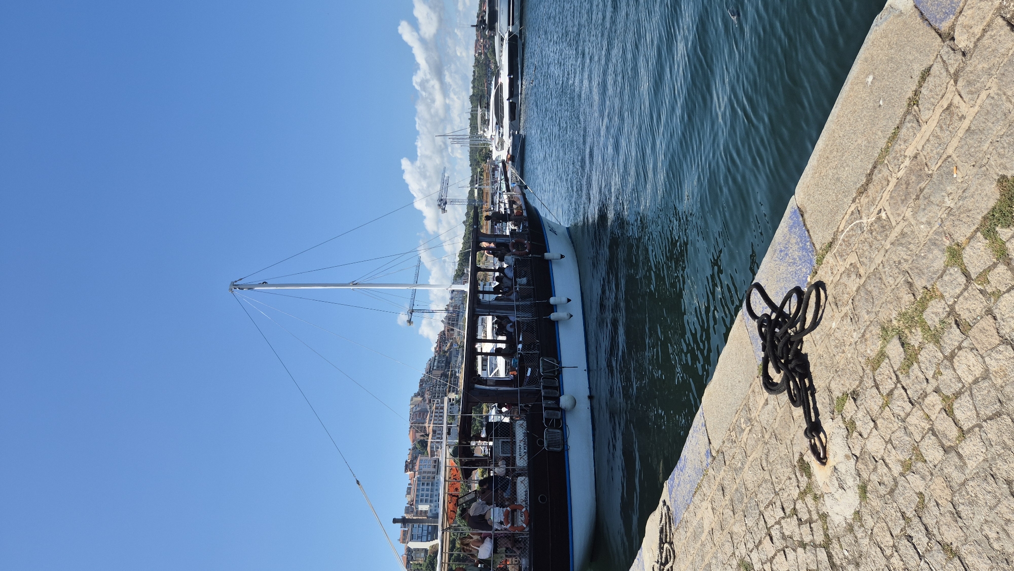 A medium-sized wooden boat with a covered passenger deck is docked in a harbor. The boat is brown with a white hull and has several passengers visible on its deck, some of whom appear to be looking out at the water. In the background, there are several white yachts, some construction cranes, and a city skyline with buildings that appear to be constructed of red-tiled roofs. The water is blue and choppy, and the sky is clear with a few white clouds. A stone walkway is in the foreground, with a thick black rope coiled on its surface. This scene is in Sozopol, Bulgaria.