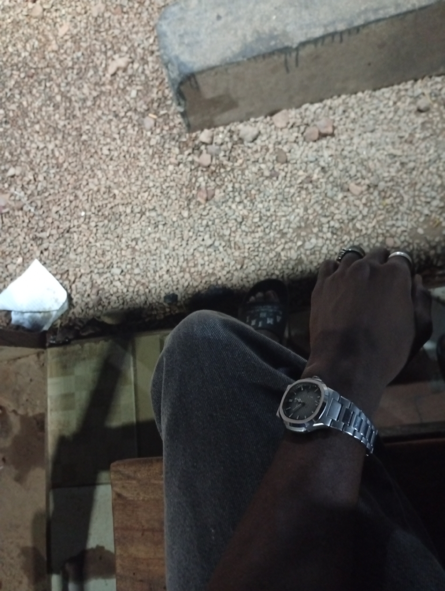 An eye-level shot depicts a person seated in an unspecified location in Adankolo, Nigeria. The individual is wearing gray jeans and a silver wristwatch on their left wrist. Their left hand, adorned with two rings, rests near their right hand, the latter also wearing a ring. Footwear in the form of sandals is visible beneath the person's leg. The surroundings include a tiled or wooden structure beneath the person, a gravelly ground surface with a concrete curb to the top and right, and a piece of white paper resting on the left-hand side.