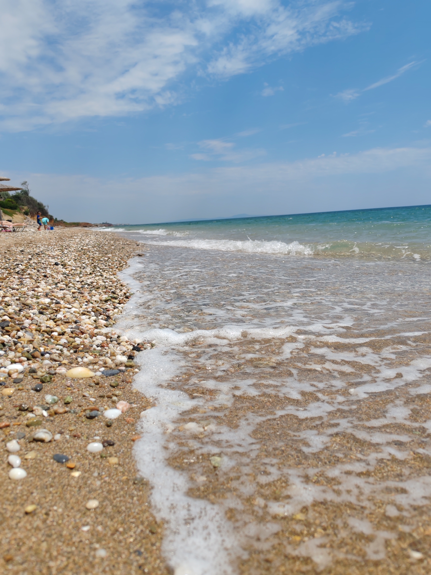 The image depicts a picturesque beach scene with a low-angle perspective. The foreground is dominated by a sandy and pebbly shoreline, with small, colorful stones scattered across the sand. Gentle waves are washing ashore, creating a foamy white surf that recedes across the beach.
In the middle ground, a few people are visible near some beach umbrellas and lounge chairs, suggesting a leisure activity. One person appears to be standing, and another might be sitting. Further back, the sea stretches out to the horizon, with a clear blue sky above dotted with white clouds. The water transitions from a turquoise hue near the shore to a deeper blue further out. The overall impression is of a sunny day at a tranquil beach location.
The time of day appears to be daytime, likely late morning or afternoon, given the bright sunlight. The weather seems pleasant and clear, ideal for a beach outing. While no specific text is visible in the image, the presence of umbrellas and lounge chairs, along with the natural beauty of the sea and sky, strongly suggests a vacation or recreational setting. The scene evokes a sense of peace and relaxation. The location is identified as Makri, Greece.