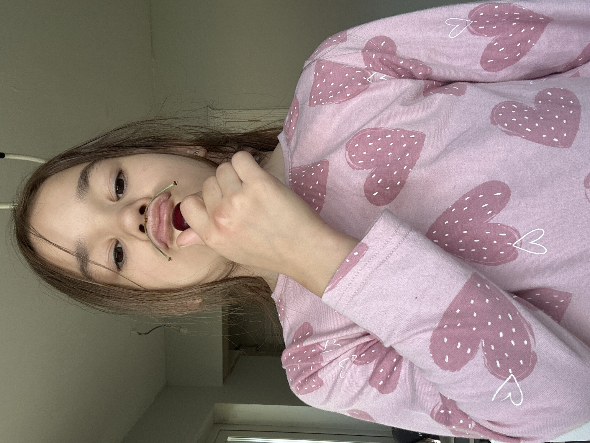 A close-up, unusually angled shot captures a young person, likely a girl, indoors in Kharnavy, Russia. The perspective is from above, looking down at her, or she is lying down looking up at the camera. She has light brown hair framing her face and is looking directly at the viewer with an open-mouthed expression. A single cherry, still attached to its stem, is held playfully between her lips. She is wearing a long-sleeved, light pink top adorned with numerous larger, darker pink heart shapes, each filled with white dots. Some of the hearts also have small, white, outlined hearts drawn on or near them. Her right hand is visible near her chin, partially obscuring her face. The background consists of a plain, light grey or off-white wall, with a window frame visible in the lower right corner, suggesting it is daytime. The overall mood is casual and playful.