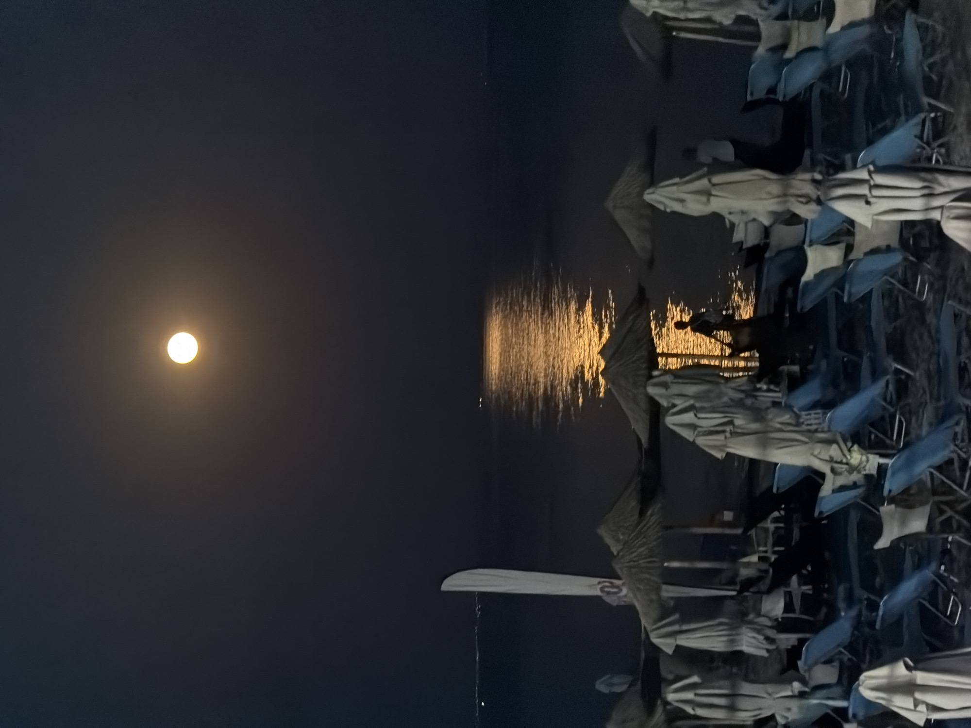 The image depicts a nighttime scene at a beach in Nea Kallikratia, Greece, illuminated by a full moon. The moon is prominently positioned in the dark sky, casting a golden reflection on the calm sea. The foreground shows a row of closed beach umbrellas and lounge chairs, suggesting the beach is unoccupied or has been prepared for the night. A white flag is visible on the left side of the image, and two faint figures are seen standing by the water's edge, silhouetted against the moonlight. The overall atmosphere is serene and tranquil.